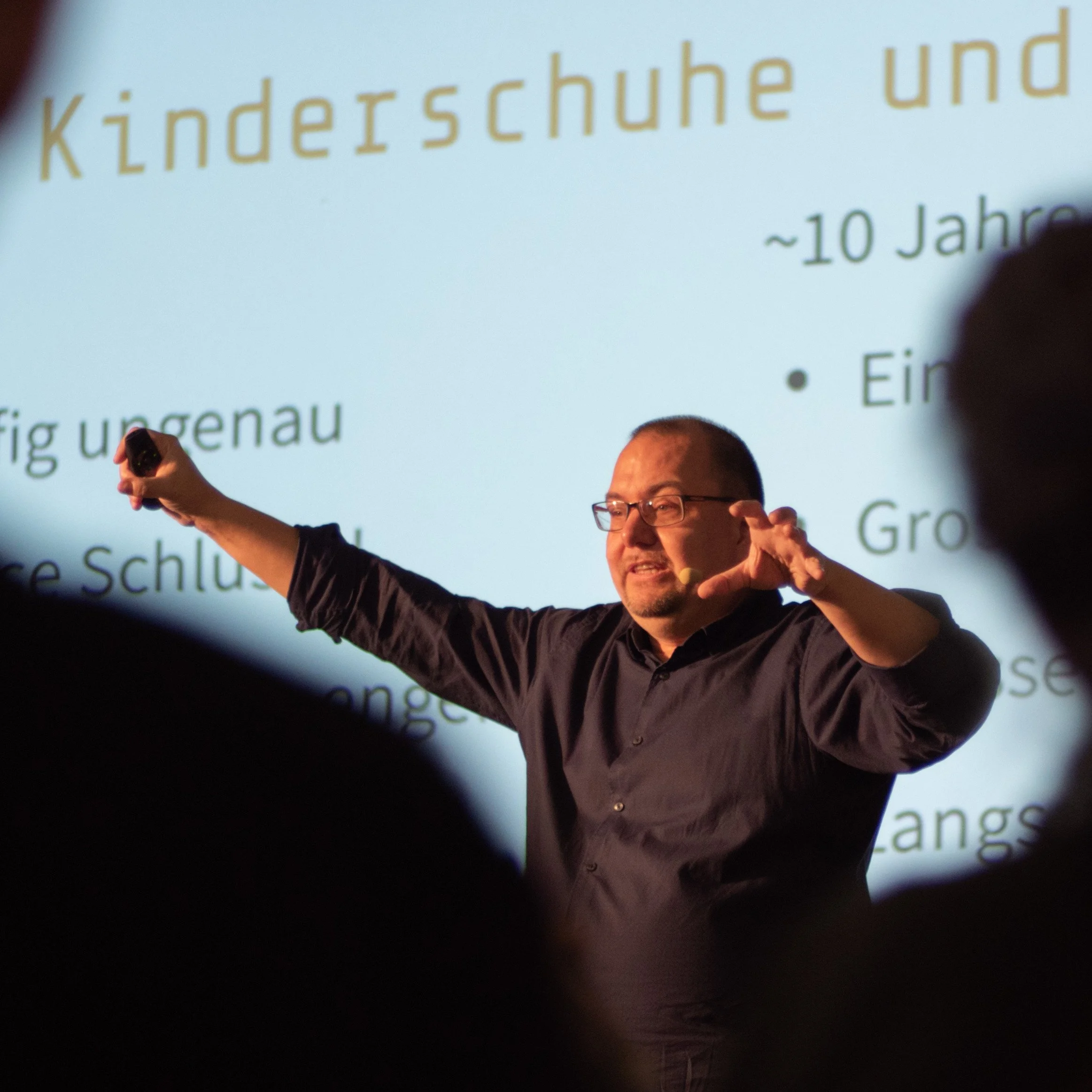 A man giving a presentation with a projected slide containing German text about children's shoes and ages, with audience members visible as blurred silhouettes in the foreground.