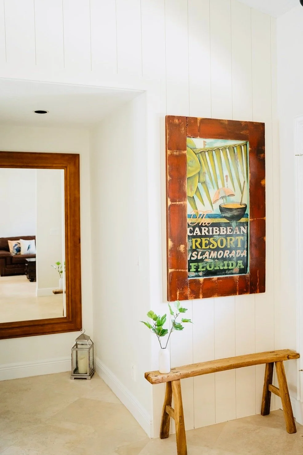 Decorative wall art with tropical theme, a small wooden bench with a white vase and green plant, and a lantern candle holder near a large mirror in a bright room.
