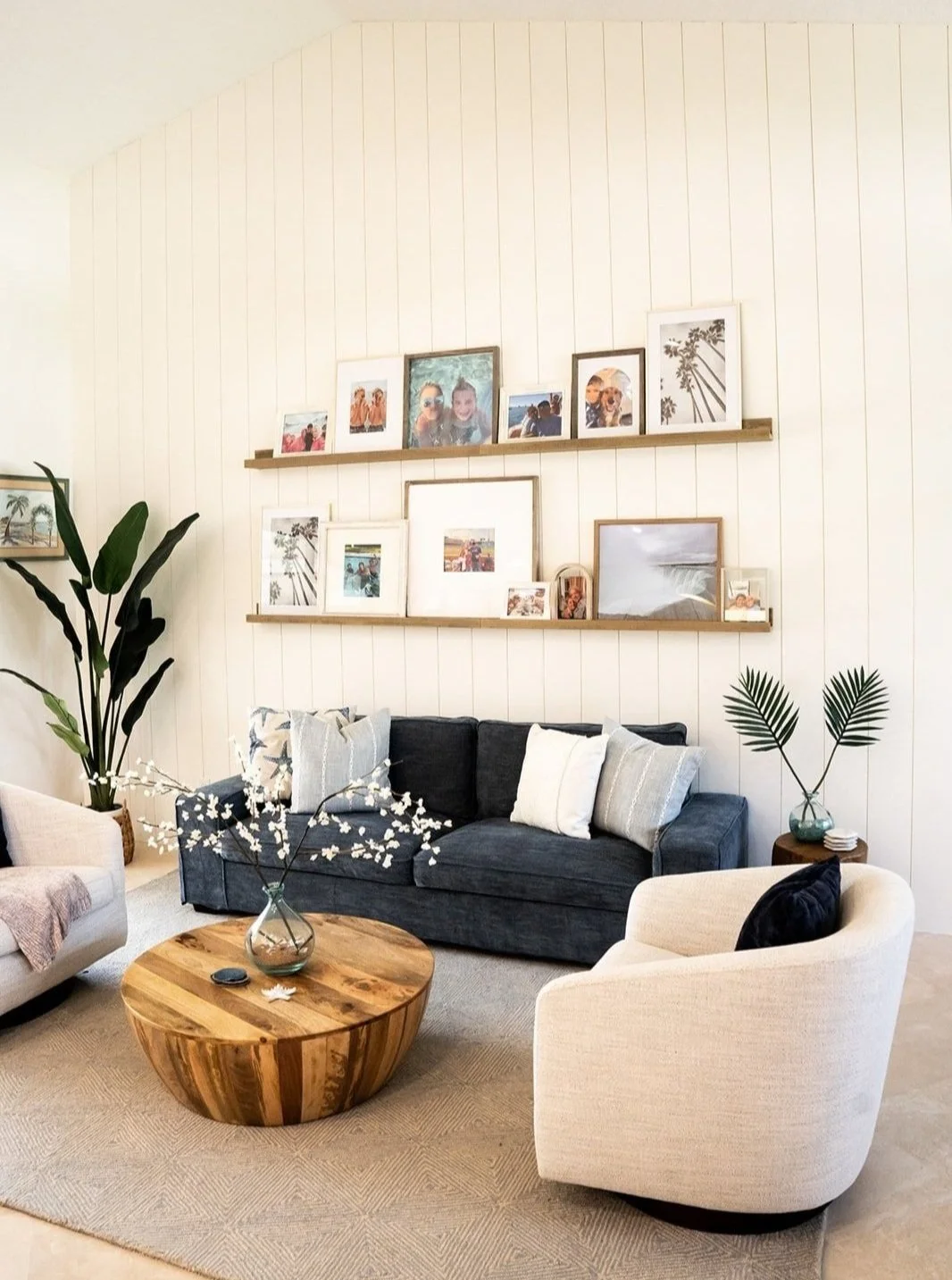 Living room with a navy blue sofa, cream armchair, round wooden coffee table, and decorative pillows. Wall with white vertical paneling and two shelves displaying framed photos and artwork. Large green plants on either side of the sofa.