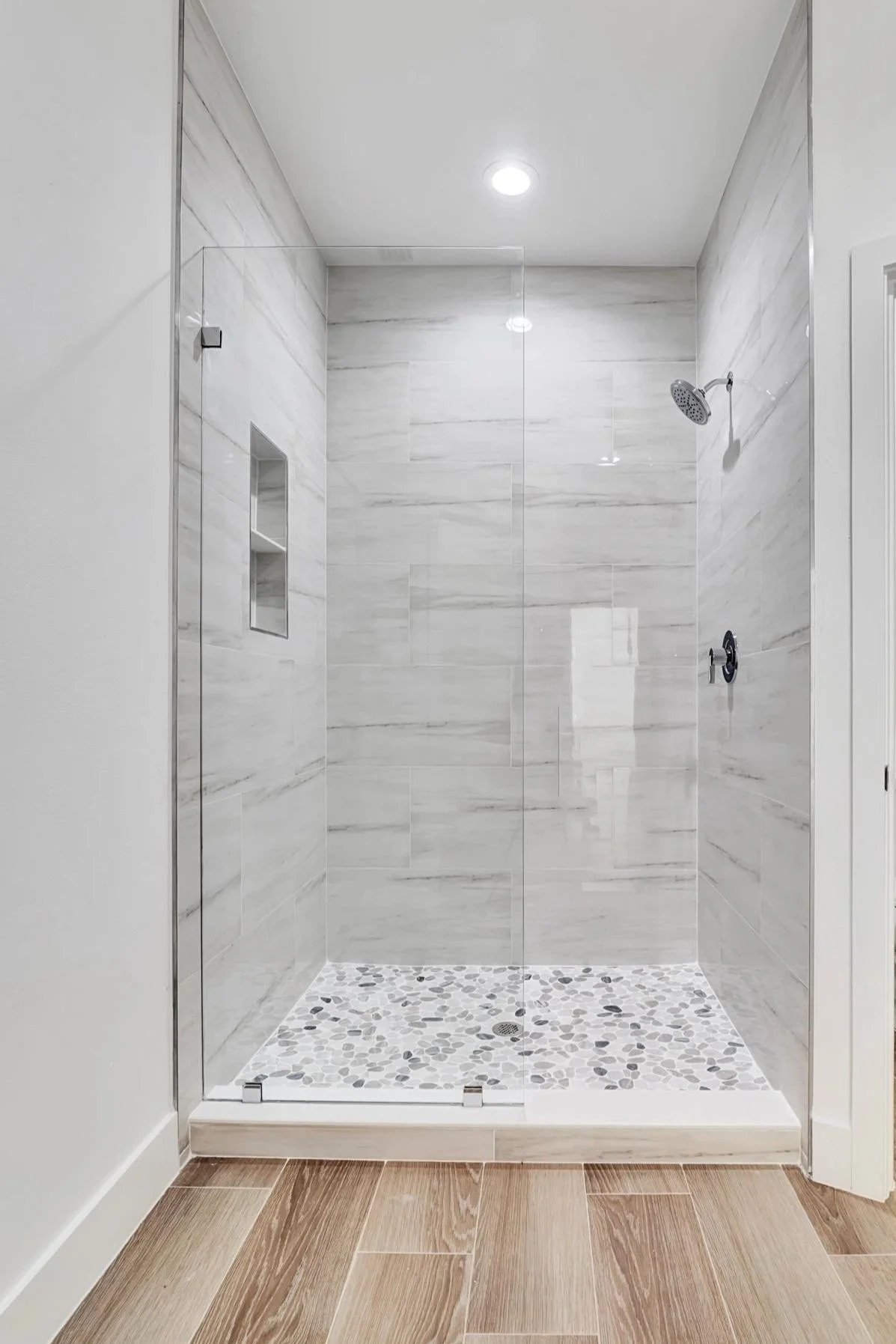 Empty walk-in shower with grey marble tiles on the walls and pebble tiles on the floor, enclosed by a clear glass door.