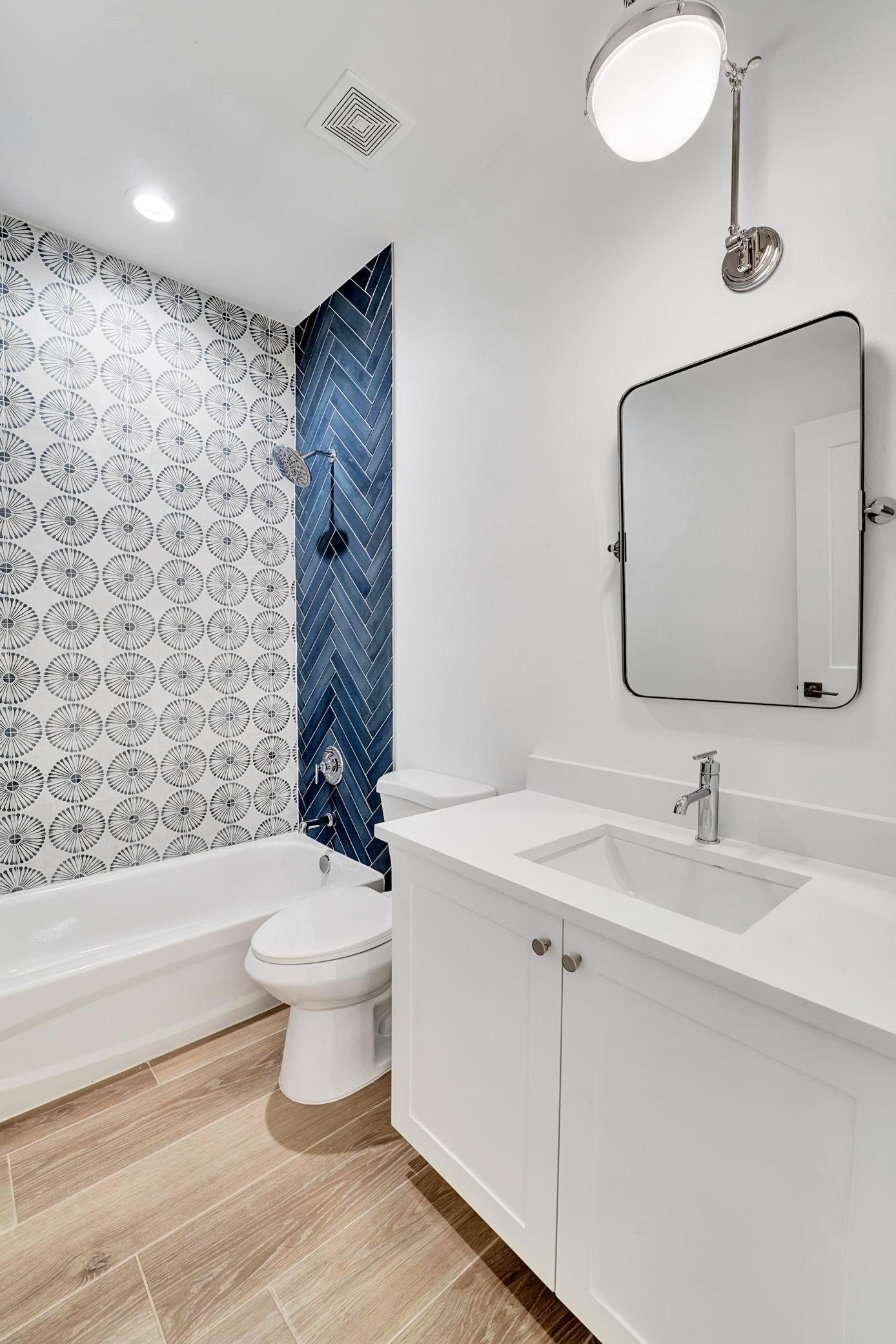 Modern bathroom with white vanity, rectangular mirror, wall-mounted light fixture, toilet, and a shower area with patterned tile including white, gray, and blue tiles.