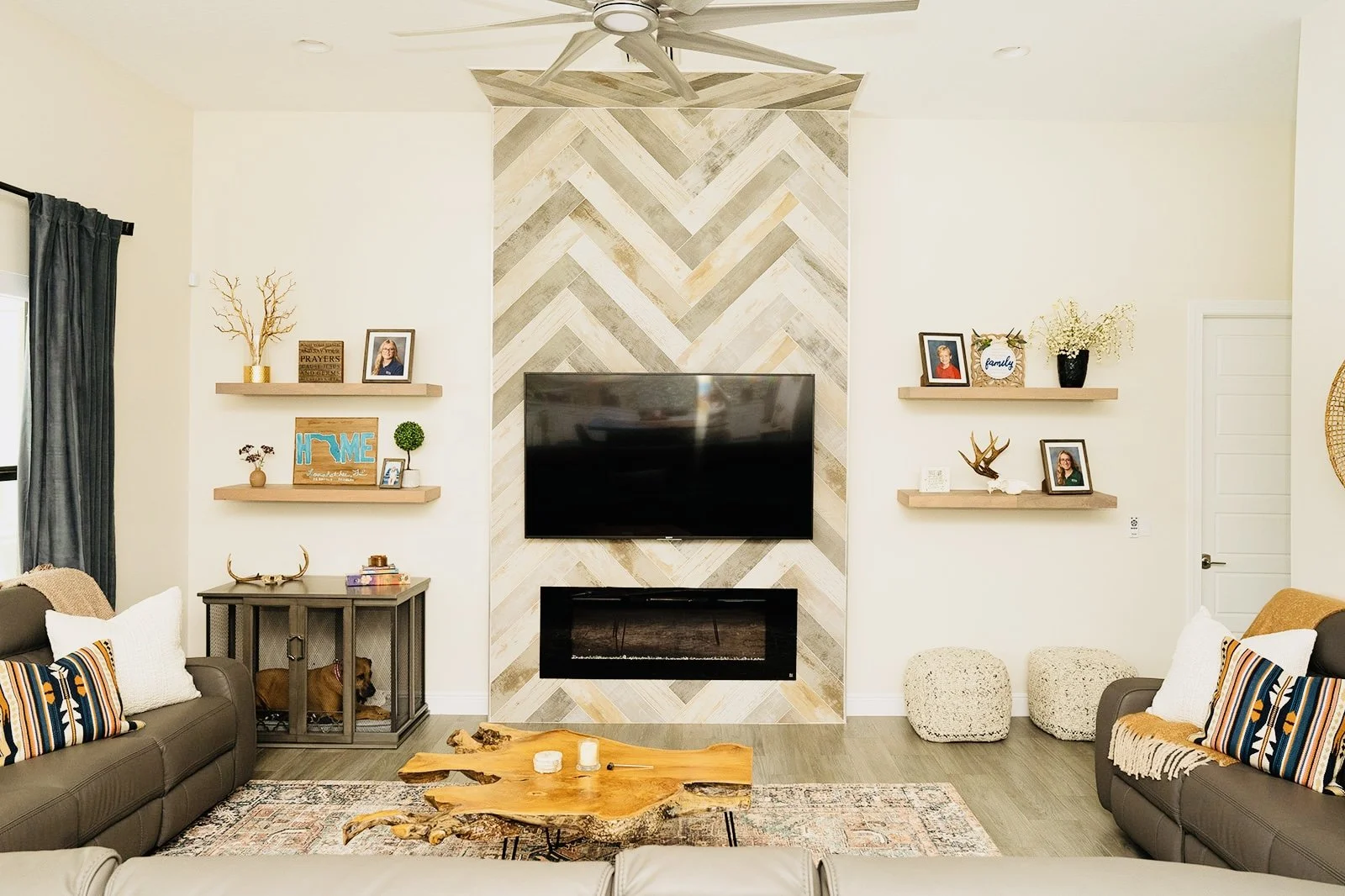 Living room with a decorated accent wall featuring a mounted flat screen TV and a fireplace, flanked by two wooden shelves with decorative items, cozy sofas with patterned pillows, a rustic wooden coffee table, and two cream-colored ottomans.