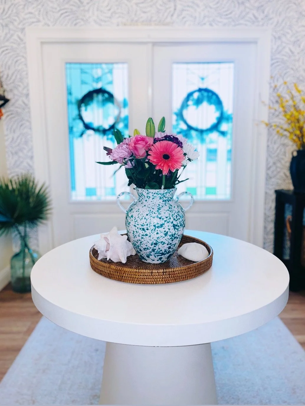 A white round table with a speckled blue and white vase filled with pink and purple flowers, placed on a woven tray with seashells, in a room with patterned wallpaper and large windows with decorative wreaths.