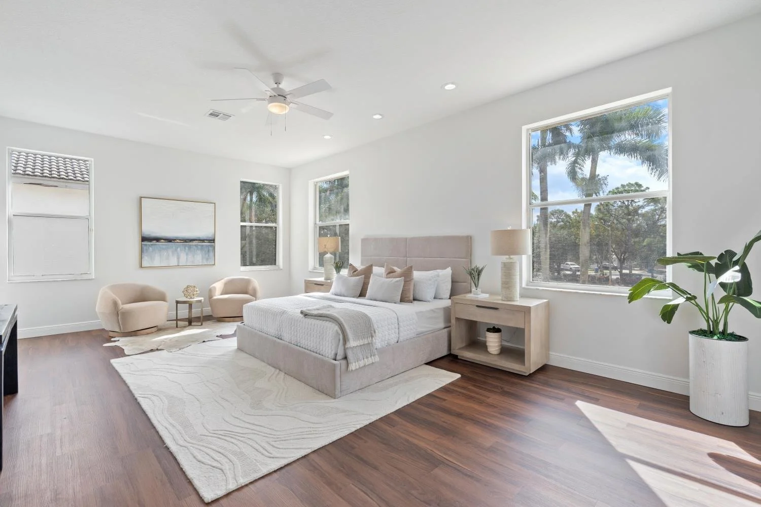 Bright bedroom with a beige bed, two nightstands with lamps, two beige armchairs, a white rug, and large windows with a view of trees.