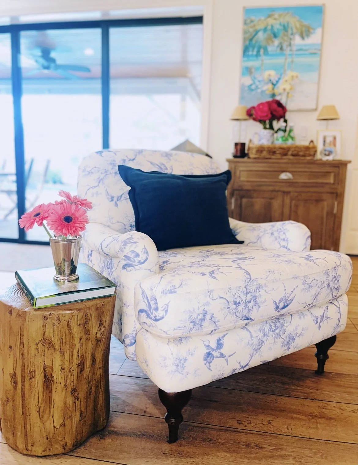 A cozy living room corner with a floral armchair, a side table with pink flowers, and a wooden cabinet with flower arrangements and paintings in the background.