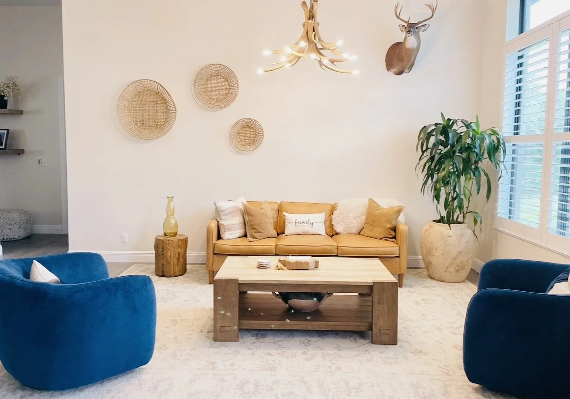 Living room with a tan leather sofa, two blue velvet armchairs, a wooden coffee table, a large potted plant, wall decor, a mounted deer head, and a chandelier made from antlers.