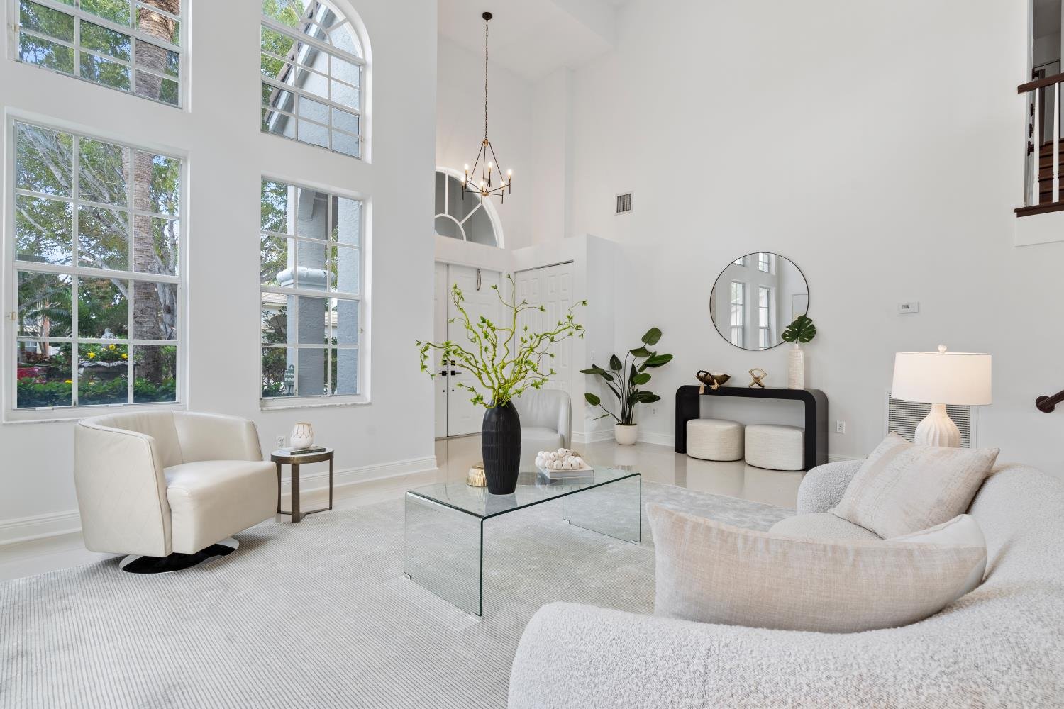 Bright living room with large arched windows, white walls, white furniture, a black vase with green branches on a glass coffee table, potted plants, and a round mirror on the wall.