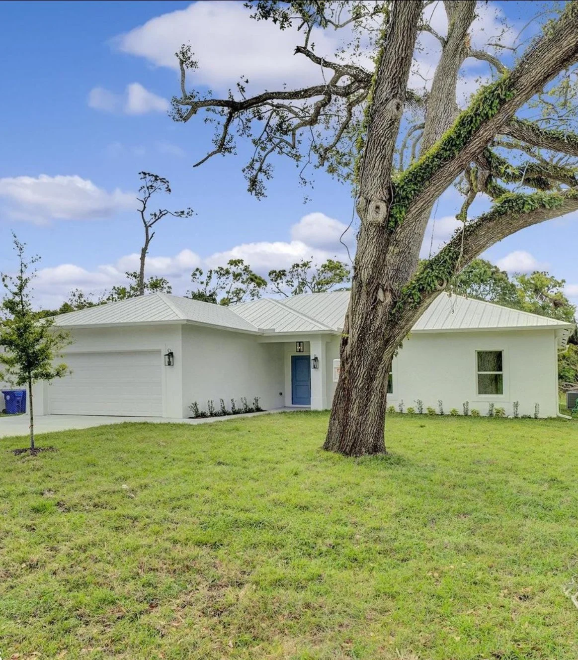 A modern white house with a blue front door, a garage, and a well-maintained lawn, featuring a large tree with a curved trunk in the front yard under a partly cloudy sky.