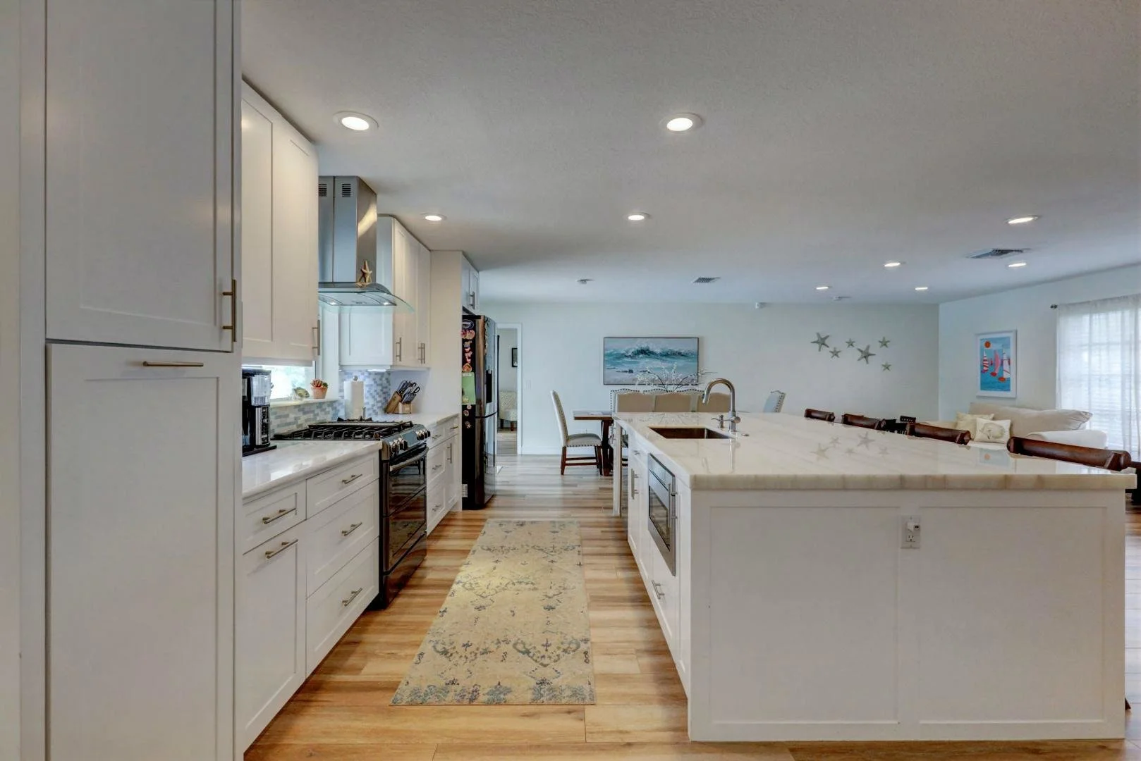 Modern kitchen with white cabinets, a large island, stainless steel appliances, a wooden floor, and a breakfast bar with chairs, open to a living and dining area.