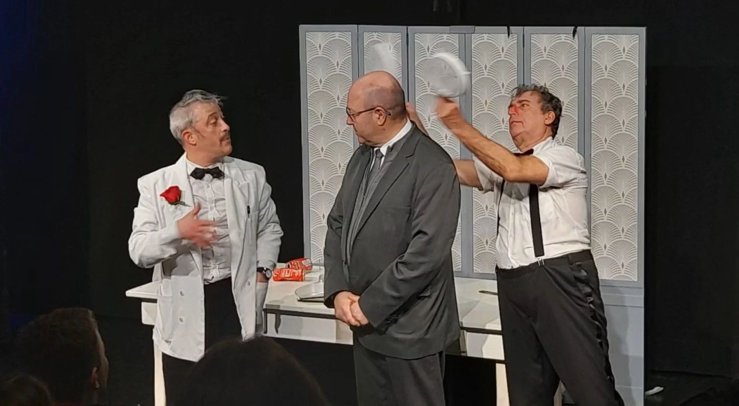 Three men are on a stage during a theatrical performance. The man on the left wears a white jacket with a red flower and a bow tie, gesturing with his hand. The man in the middle is dressed in a gray suit, looking downward. The man on the right, in a white shirt and suspenders, is swinging a plate. A white table with items and a decorative screen are in the background.