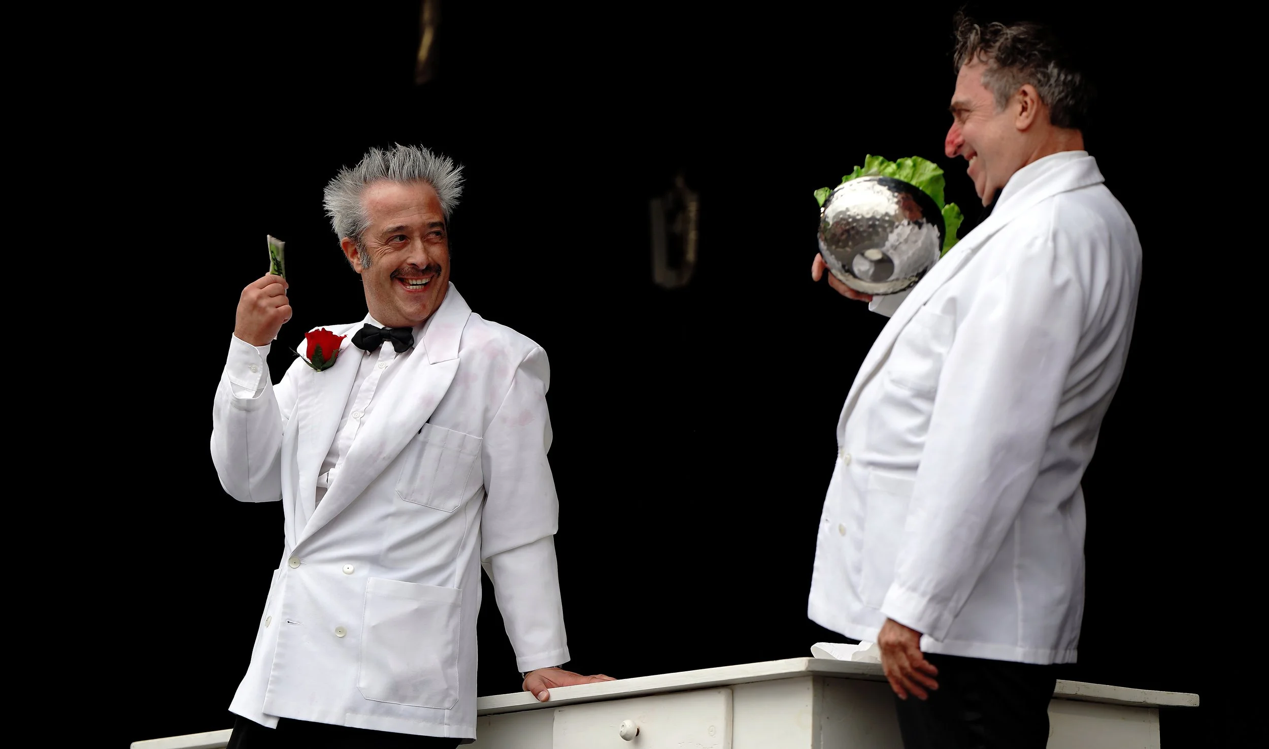 Two men dressed as waiters in white tuxedos with black bow ties, smiling and greeting each other, with one holding a dollar bill and the other holding a salad in a silver bowl, against a black background.