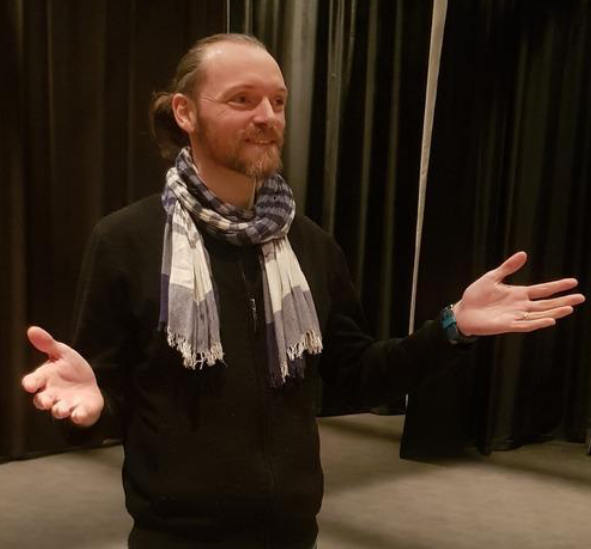 A man with long hair and a beard, wearing a black jacket and a striped scarf, standing with his arms outstretched in front of black curtains, smiling.