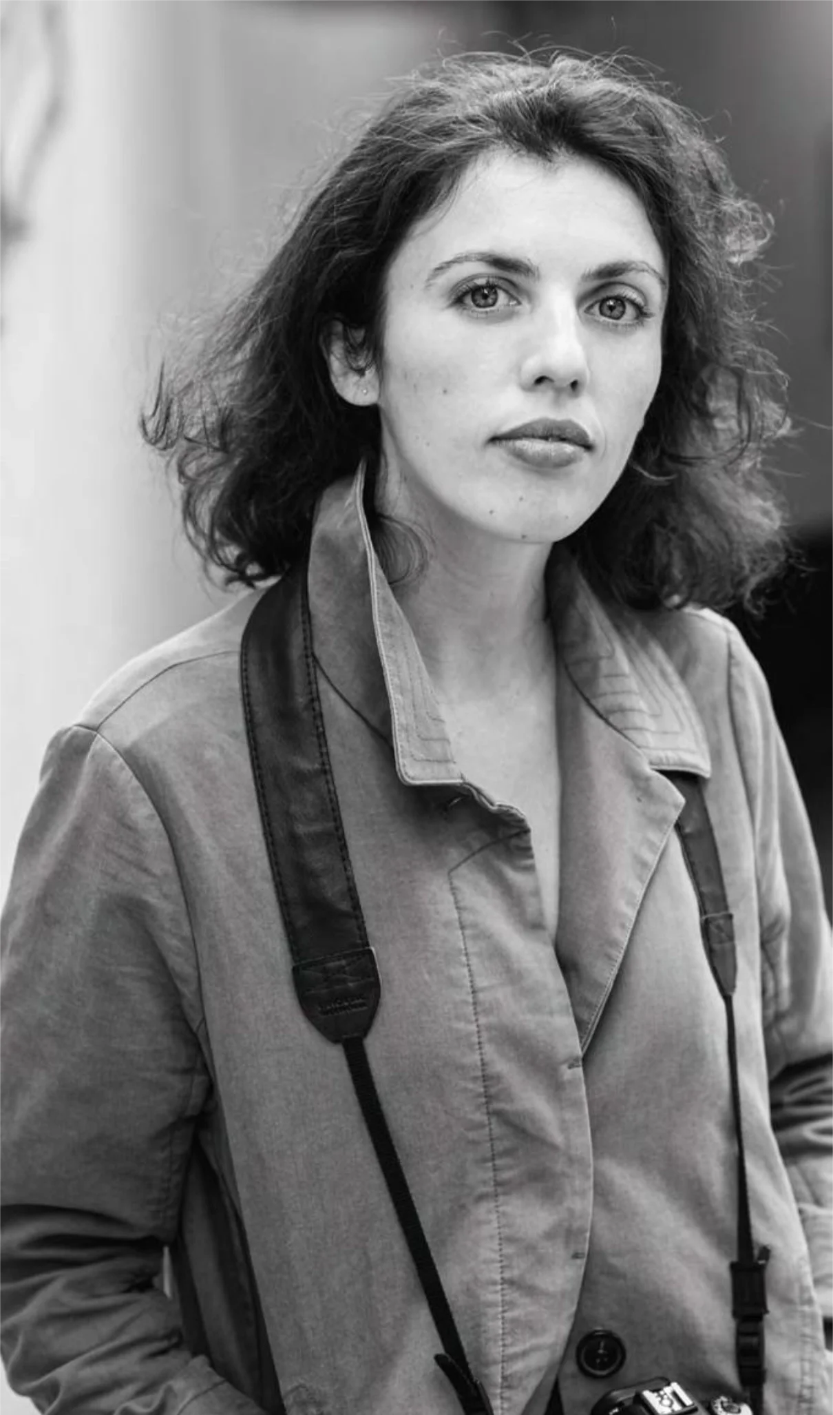 Black and white photograph of a woman with shoulder-length curly hair, wearing a collared jacket, with a camera hanging from her neck.