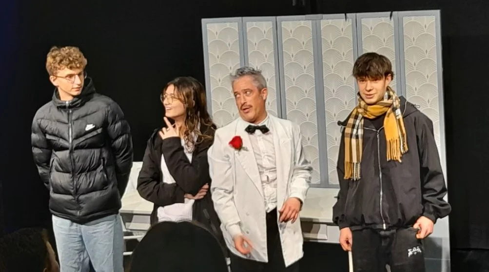 Four young people standing on a stage with a white classical-style backdrop, including two men, one dressed as a chef, and two women, dressed casually, engaged in a performance or play.