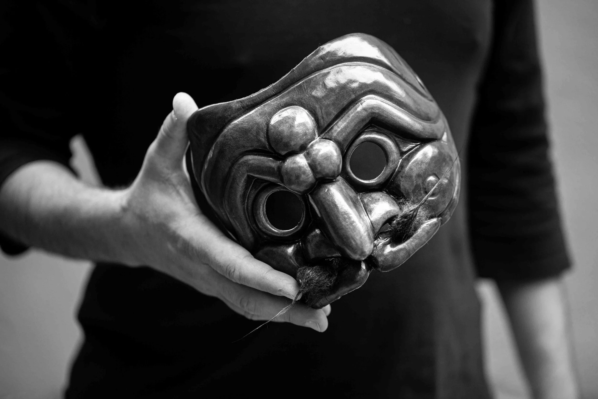 Person holding a carved mask with exaggerated facial features and holes for eyes, made of dark material.