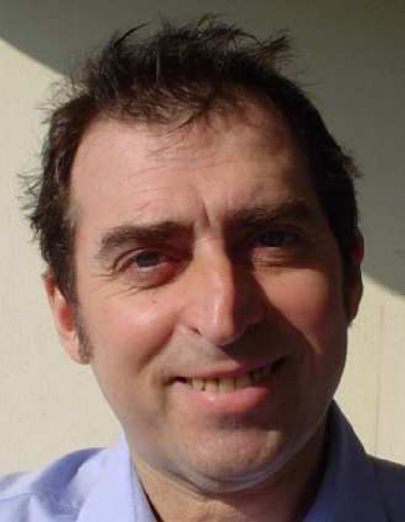 Close-up of a man's face with messy dark hair, smiling slightly, wearing a light blue collared shirt.