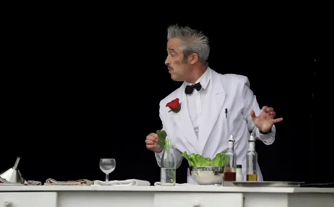 A man dressed as a chef in a white coat and bow tie, holding a red rose and gesturing with his right hand, standing behind a table with various bottles, a glass, and a bowl of greens, against a black background.