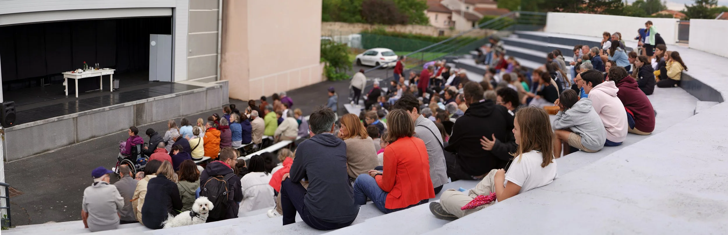 An outdoor auditorium with an audience watching a stage with tables, bottles, and glasses. The audience is sitting on tiered concrete steps and includes people, children, and a dog, with some wearing casual clothing and jackets. The background features a residential area with parked cars.
