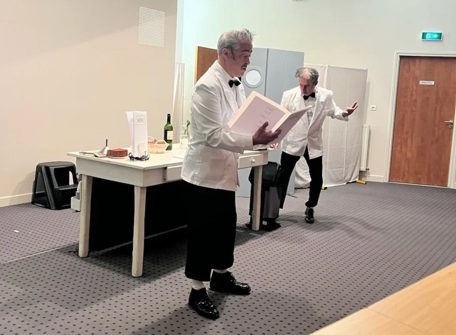 Two men dressed as waiters or performers in tuxedos with tails are standing in a room with plain white walls and a gray carpeted floor. One is reading from a large book or menu, and the other is gesturing with his right hand. There is a white table with a bottle of wine, a cake, plates, and other items, along with two black stools. A door and a bright emergency exit sign are visible on the wall.