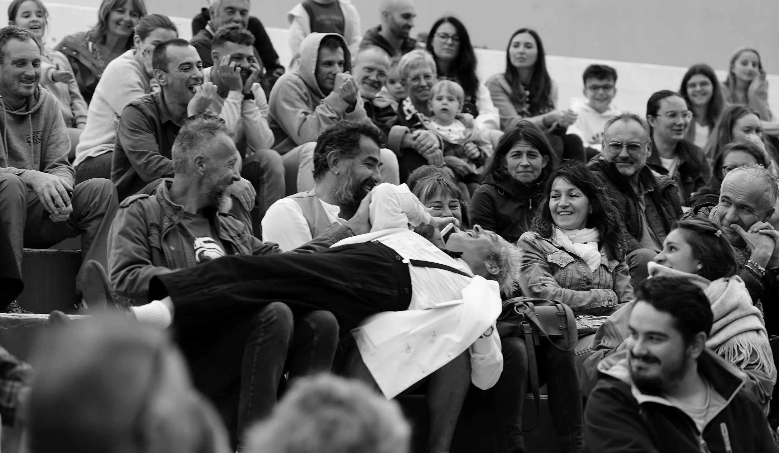 A group of people sitting in an auditorium, smiling and laughing, with a man lying across some seats, holding and looking at a baby.