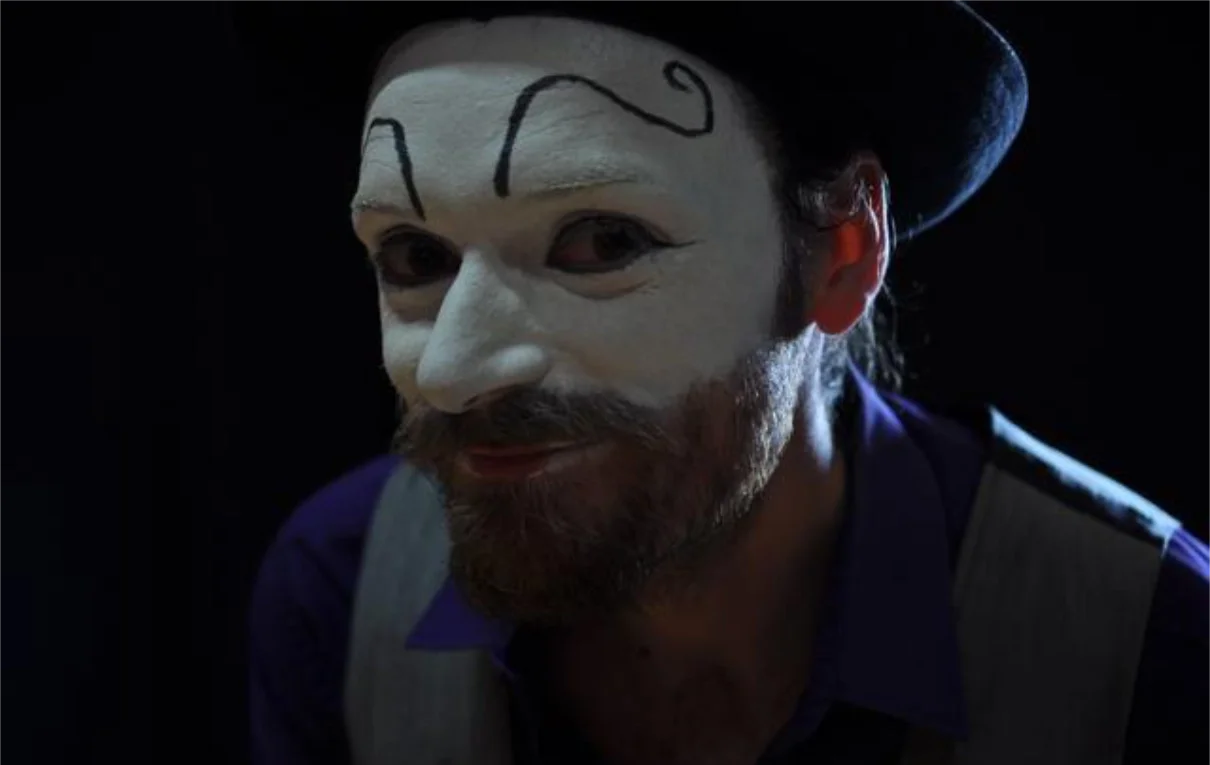 A person wearing a white painted face with black designs, a dark hat, and a purple shirt, possibly in costume or makeup for a performance.
