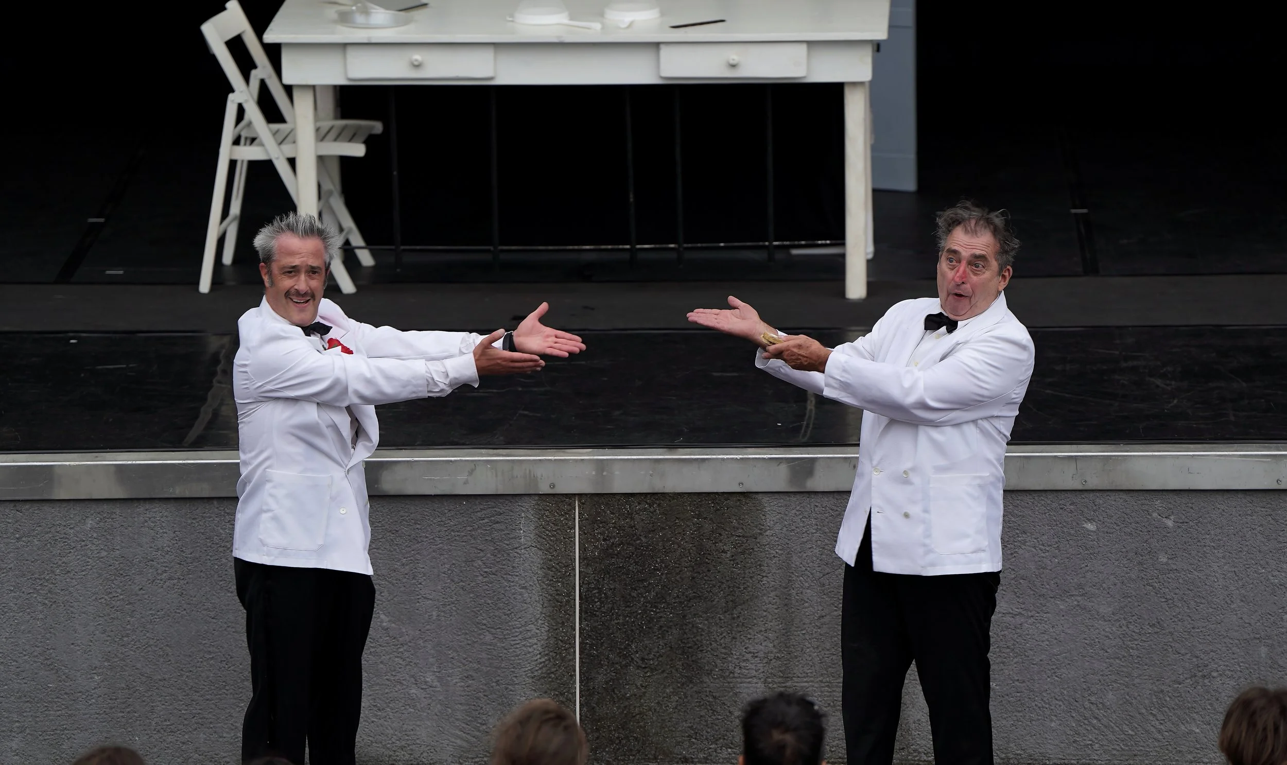 Two men dressed as waiters in white jackets and black bow ties are performing a theatrical act on stage, gesturing towards each other with open hands, in front of a black and gray stage backdrop.