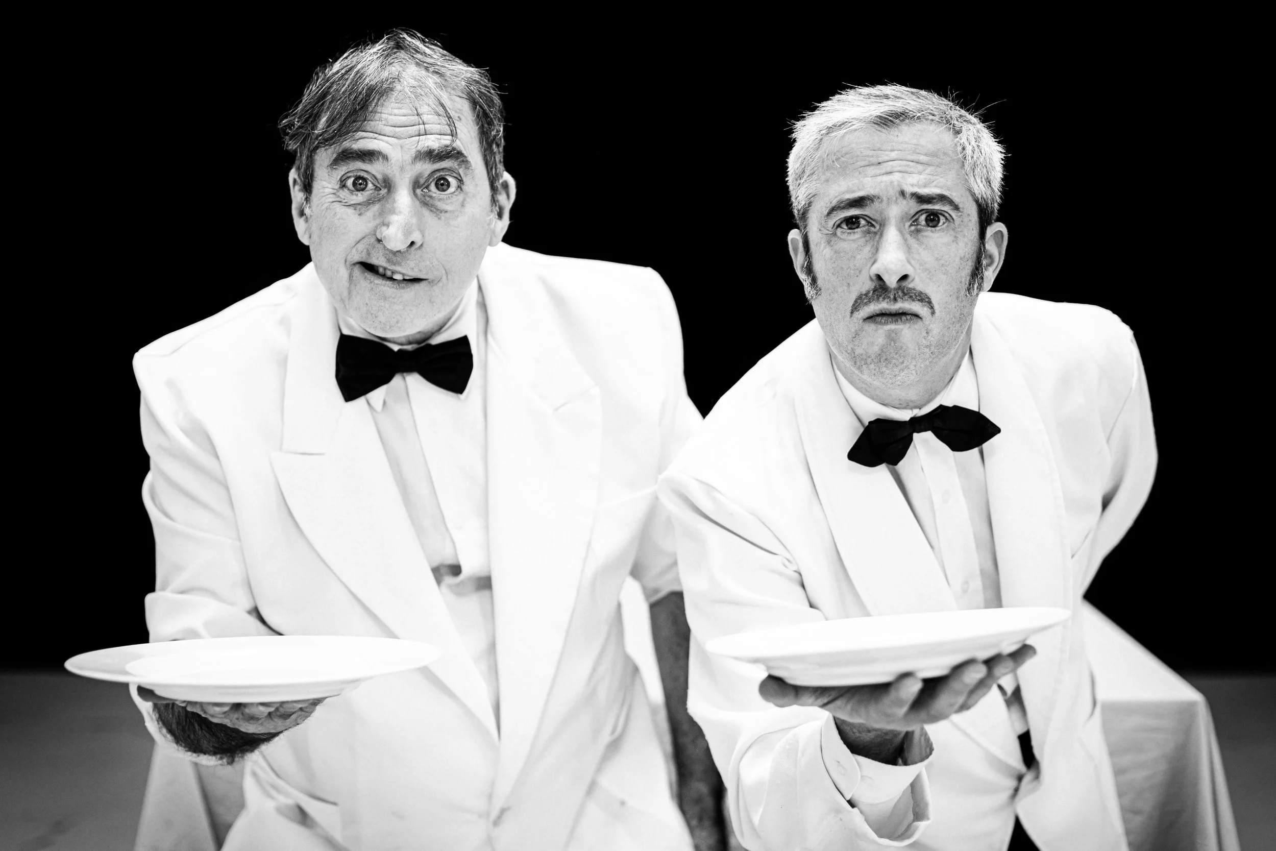 Two men dressed as waiters in white suits with black bow ties, holding empty plates and looking at the camera with comedic expressions, against a black background.