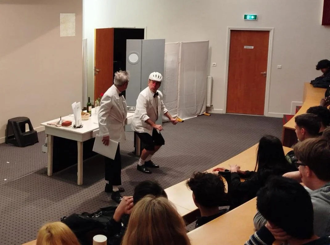 Two performers in white coats are on stage; one person is wearing a helmet and making a dance move, while the other stands nearby. They are entertaining an audience seated in a semi-circular arrangement in a classroom or lecture hall setting.