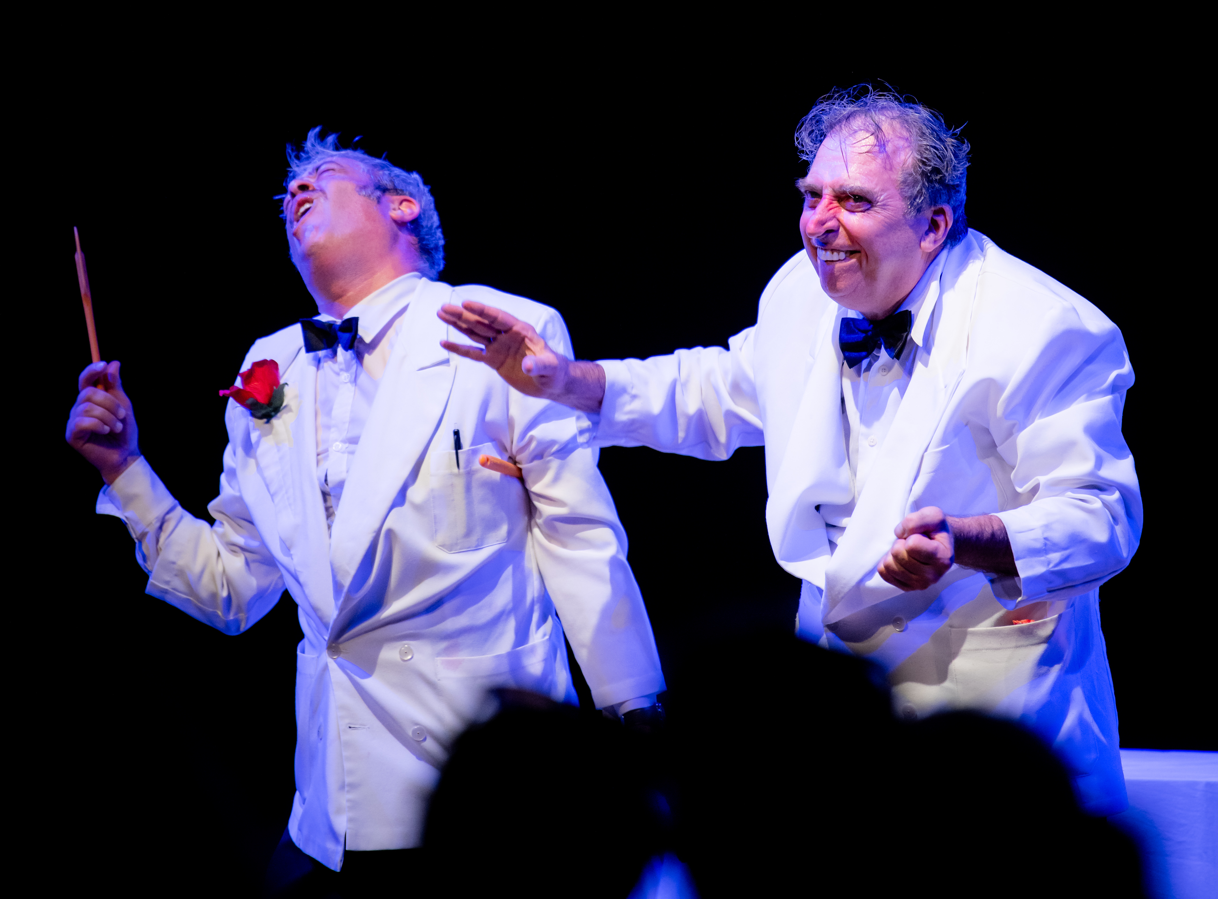 Two men dressed in white tuxedos with black bow ties, performing on stage. The man on the left holds a conductor's baton and is singing passionately, while the man on the right is smiling and gesture with his hand. Both appear to be engaged in a theatrical or musical performance.