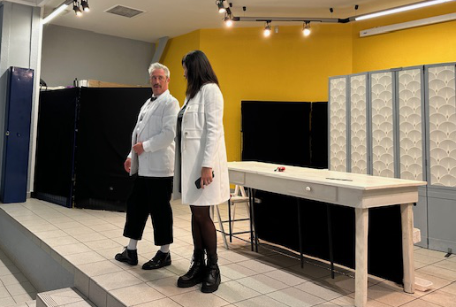 Two people wearing white lab coats standing in a room with yellow and gray walls, a large black monitor, a white table, and a telephone.