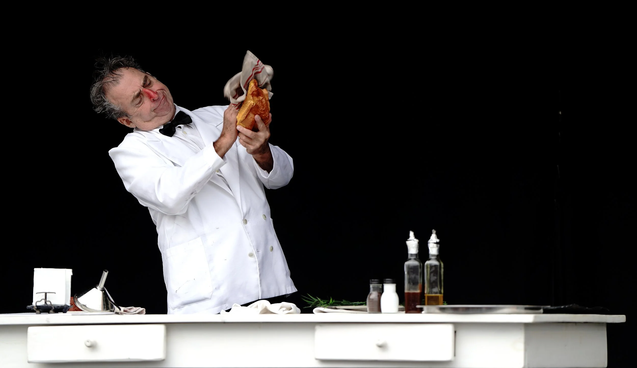 A man in a white chef's coat and black bow tie is holding a piece of bread with a hot dog in it, grimacing as if in pain, while tearing a white and red checkered cloth. The background is black, and on the white countertop in front of him are bottles, spices, and a paper towel.