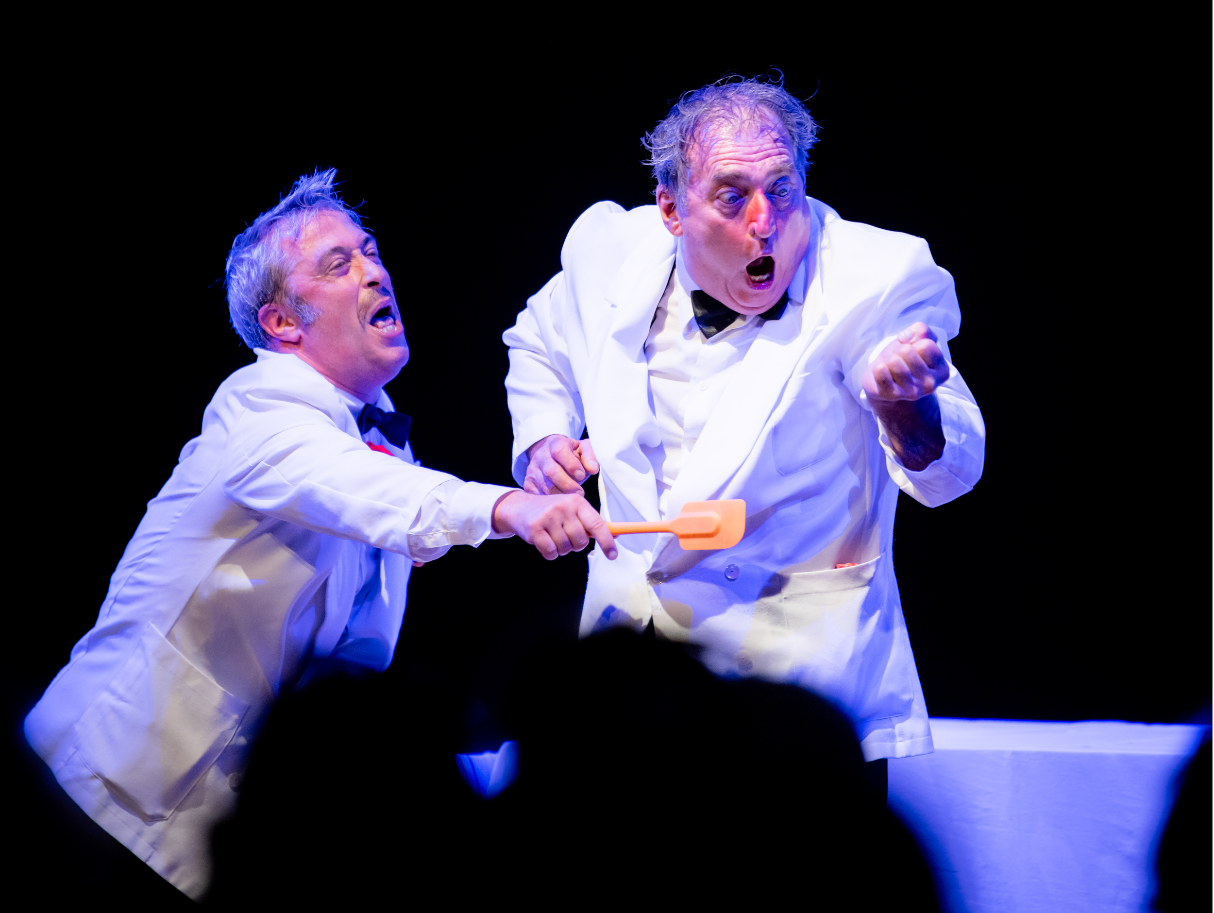 Two men dressed as chefs with white jackets and black bow ties engaged in a theatrical performance; one is grabbing the other's lapel with a distressed expression, while the other appears angry or shouting, holding a spatula.
