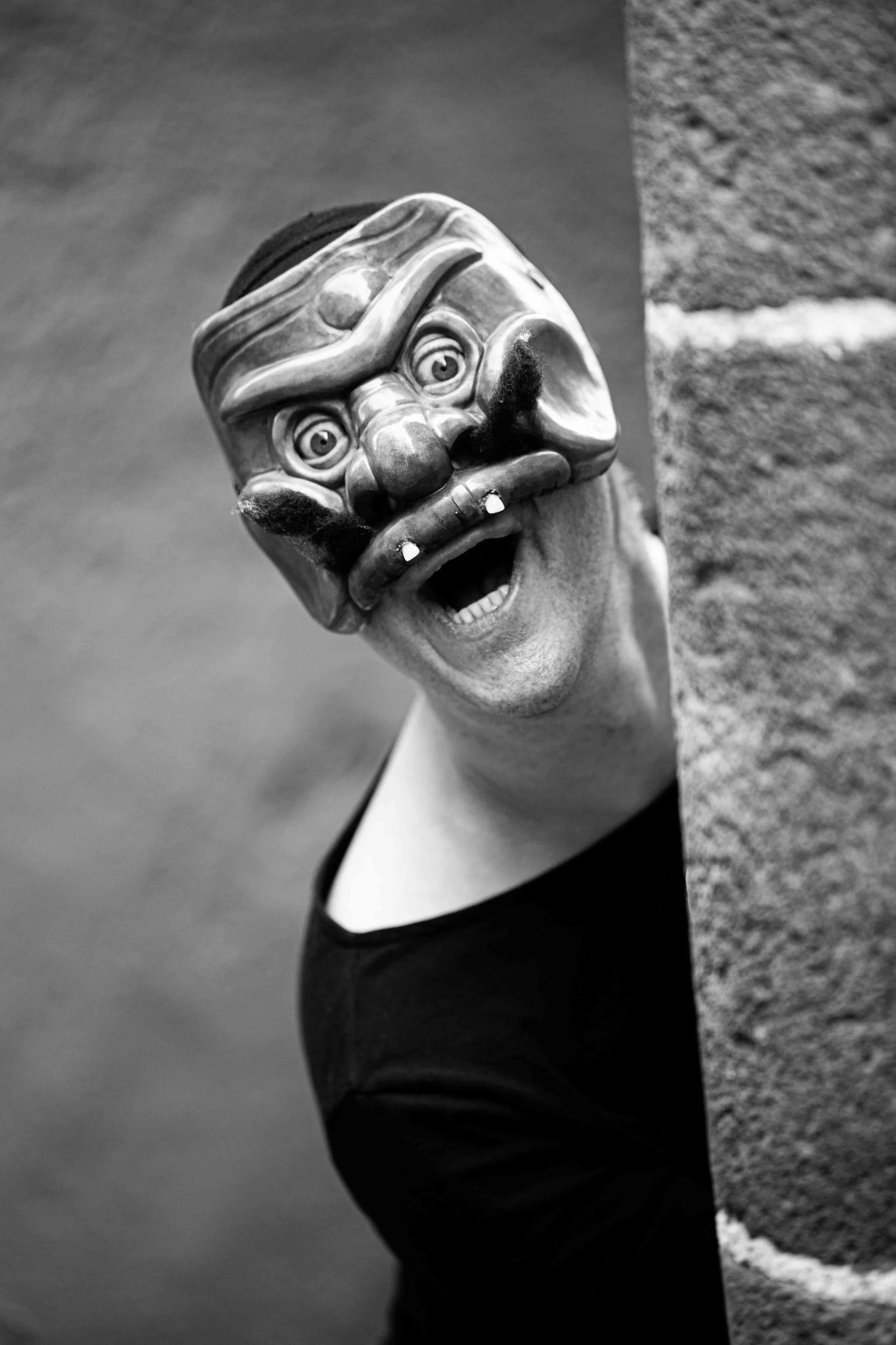 Person peeking around a corner wearing a clown mask with exaggerated facial features in black and white.