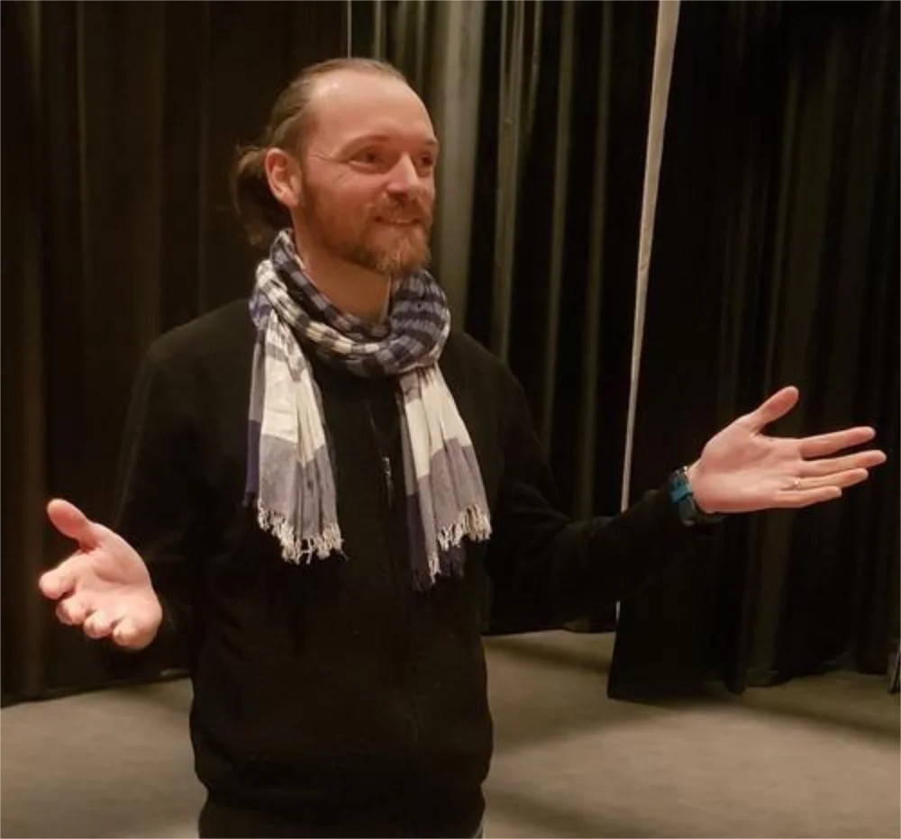 A man with long hair, a beard, and a scarf stands in front of black curtains, smiling with arms out to the sides.