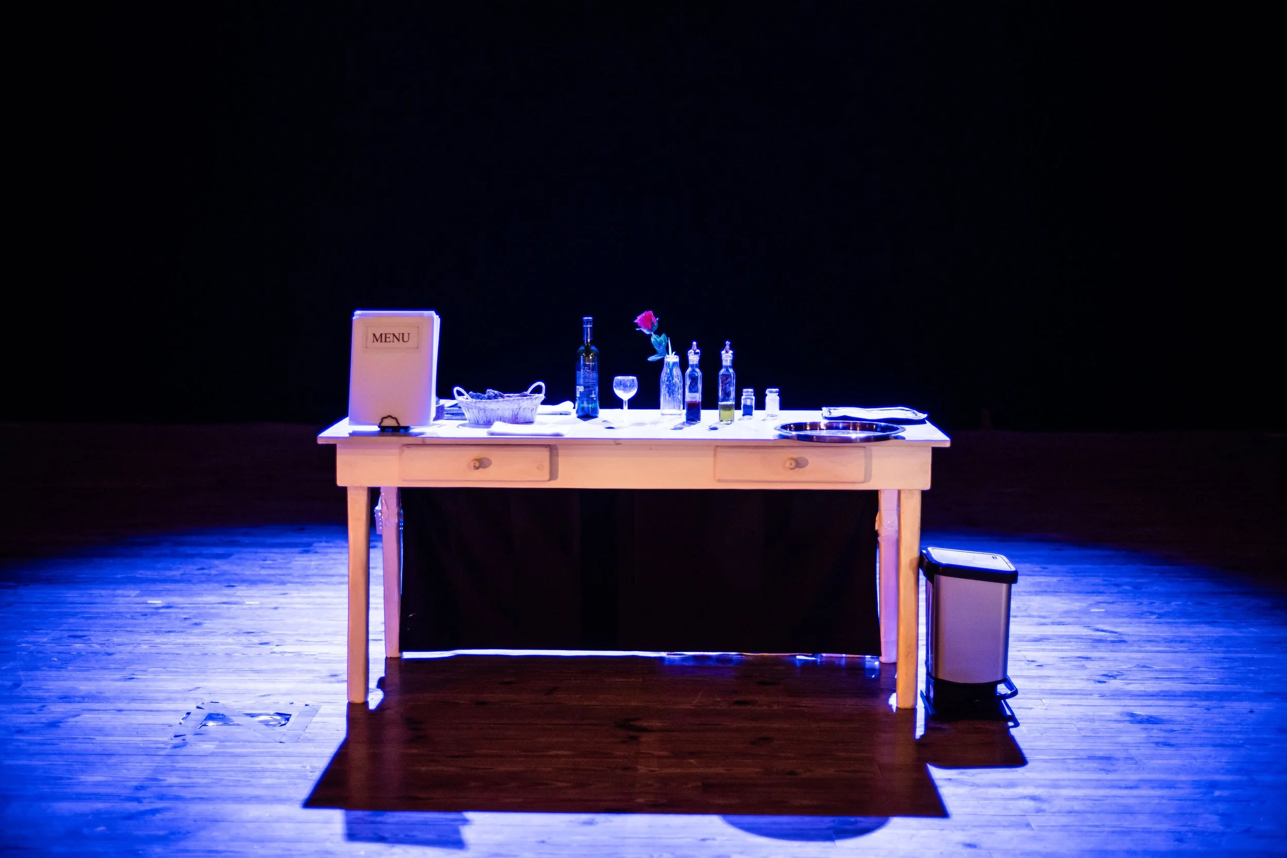 Empty table with various bottles, glasses, a menu, and a rose in a vase, lit by blue lights against a dark background.