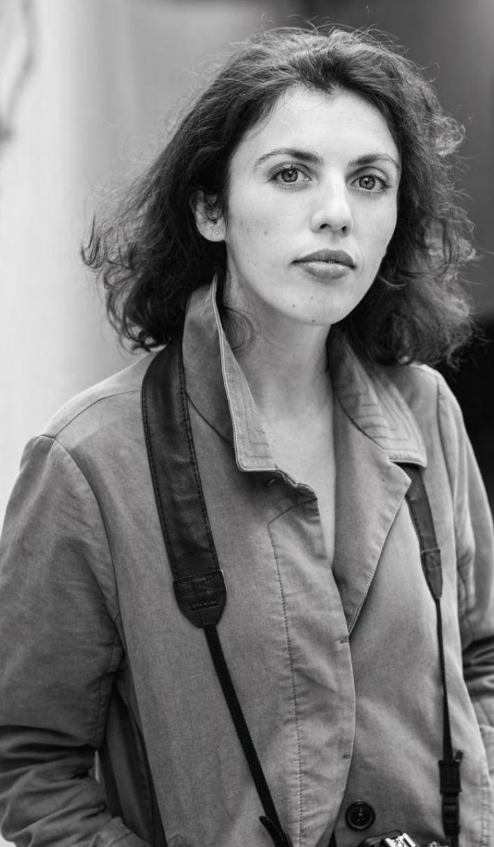 Black and white photo of a woman with wavy hair wearing a jacket and a camera strap around her neck.
