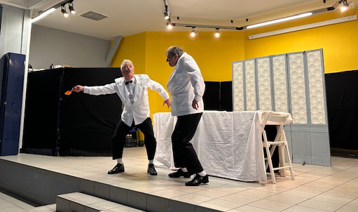 Two men in white jackets performing a comedy act on stage with a table and folding screen in the background.
