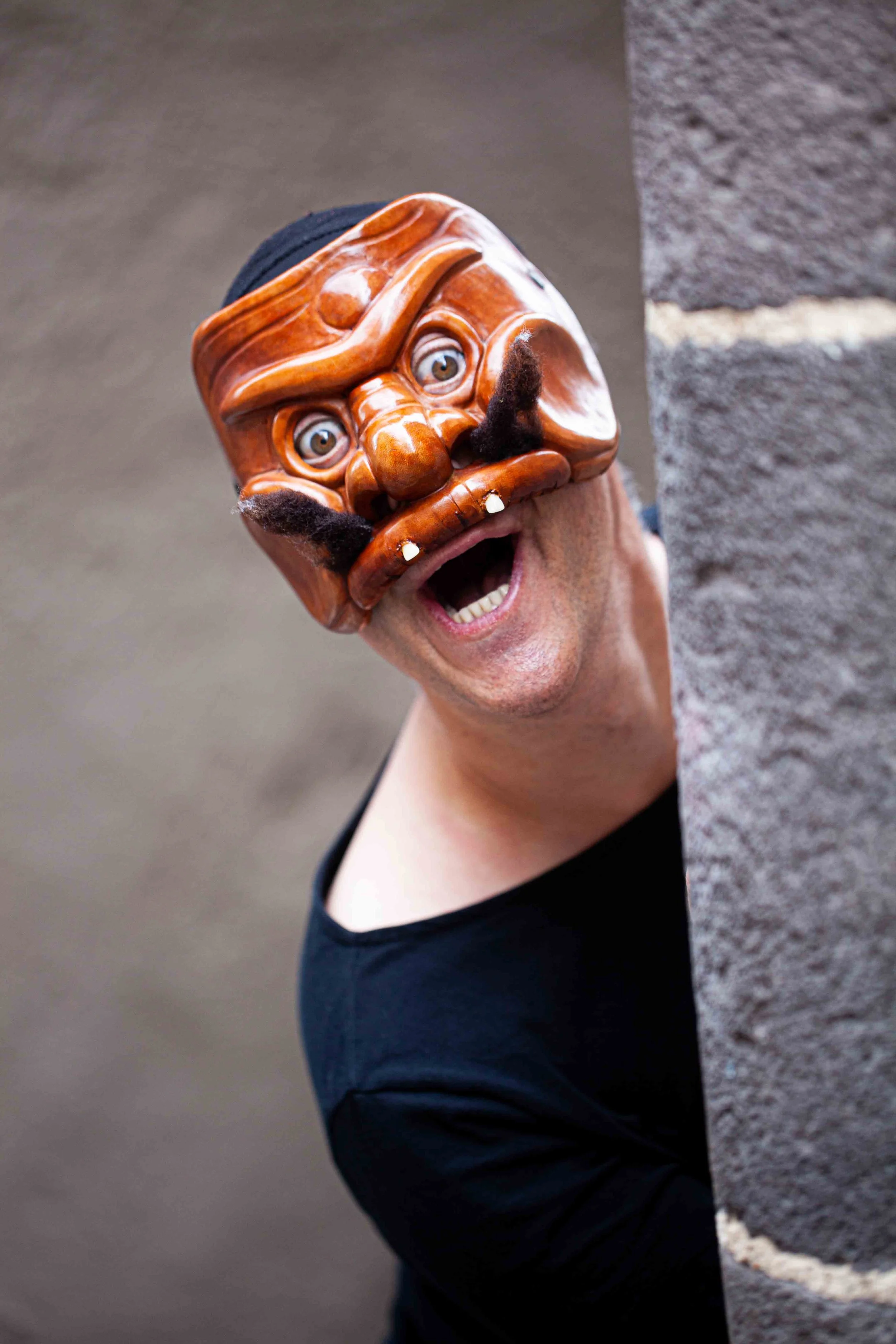 A person wearing a wooden mask with painted eyes, a mustache, and an eye patch, peeking from behind a stone wall and smiling.