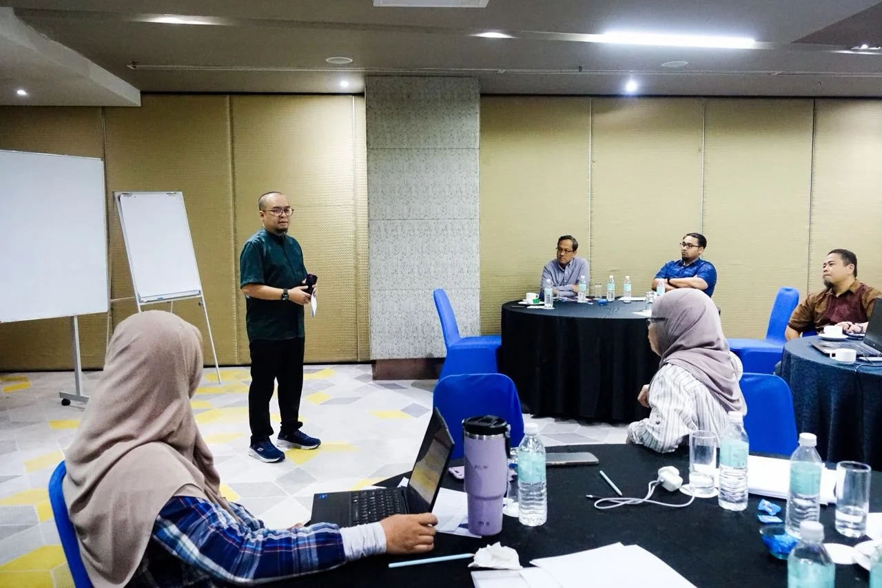 A man standing and speaking to a group of seated people in a conference room. Some people are using laptops, and there are water bottles and notebooks on the tables.