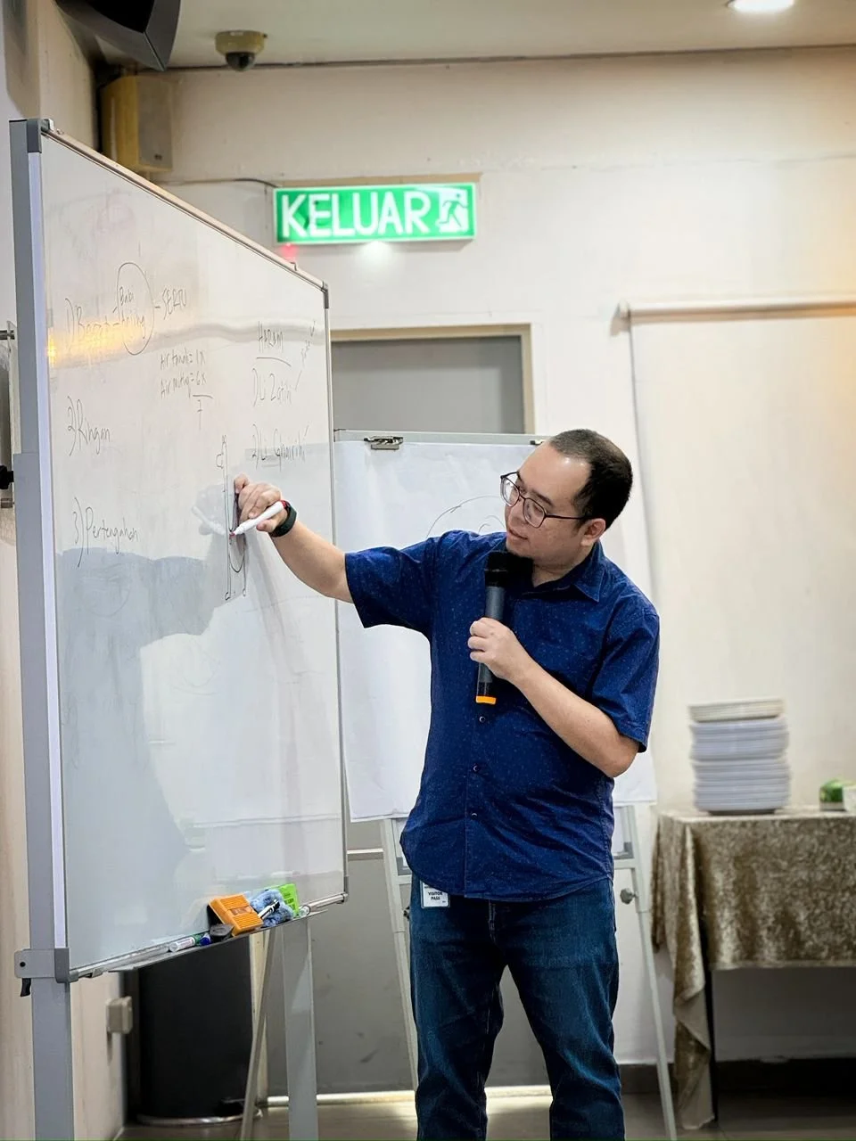 A man standing in front of a whiteboard, writing with a marker and holding a microphone, appears to be giving a presentation or lecture. There is a stack of plates on a table in the background and an illuminated exit sign labeled 'KELUAR' above a doo