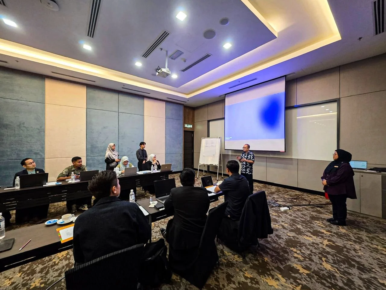 A group of people attending a business meeting or conference in a modern conference room with a large projection screen, some seated around a table with laptops, and a speaker standing in front of the room.