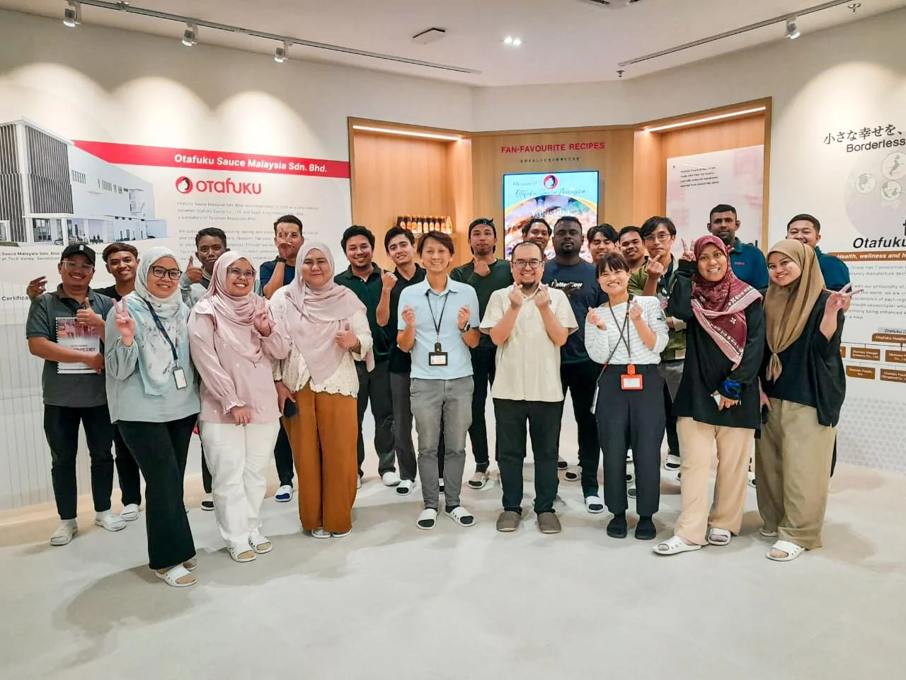 Group of diverse people standing together indoors, smiling, some making peace signs, in front of an exhibit about Otafuku sauce Malaysia.