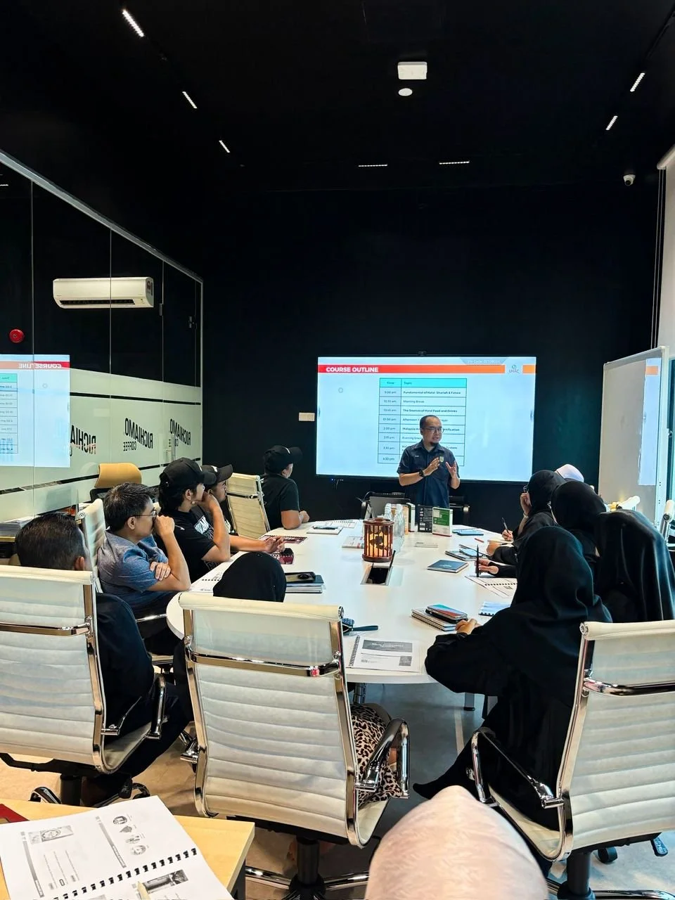 A group of people seated around a conference table in a modern, dark-colored conference room, listening to a presenter standing in front of a large digital screen displaying a course outline.
