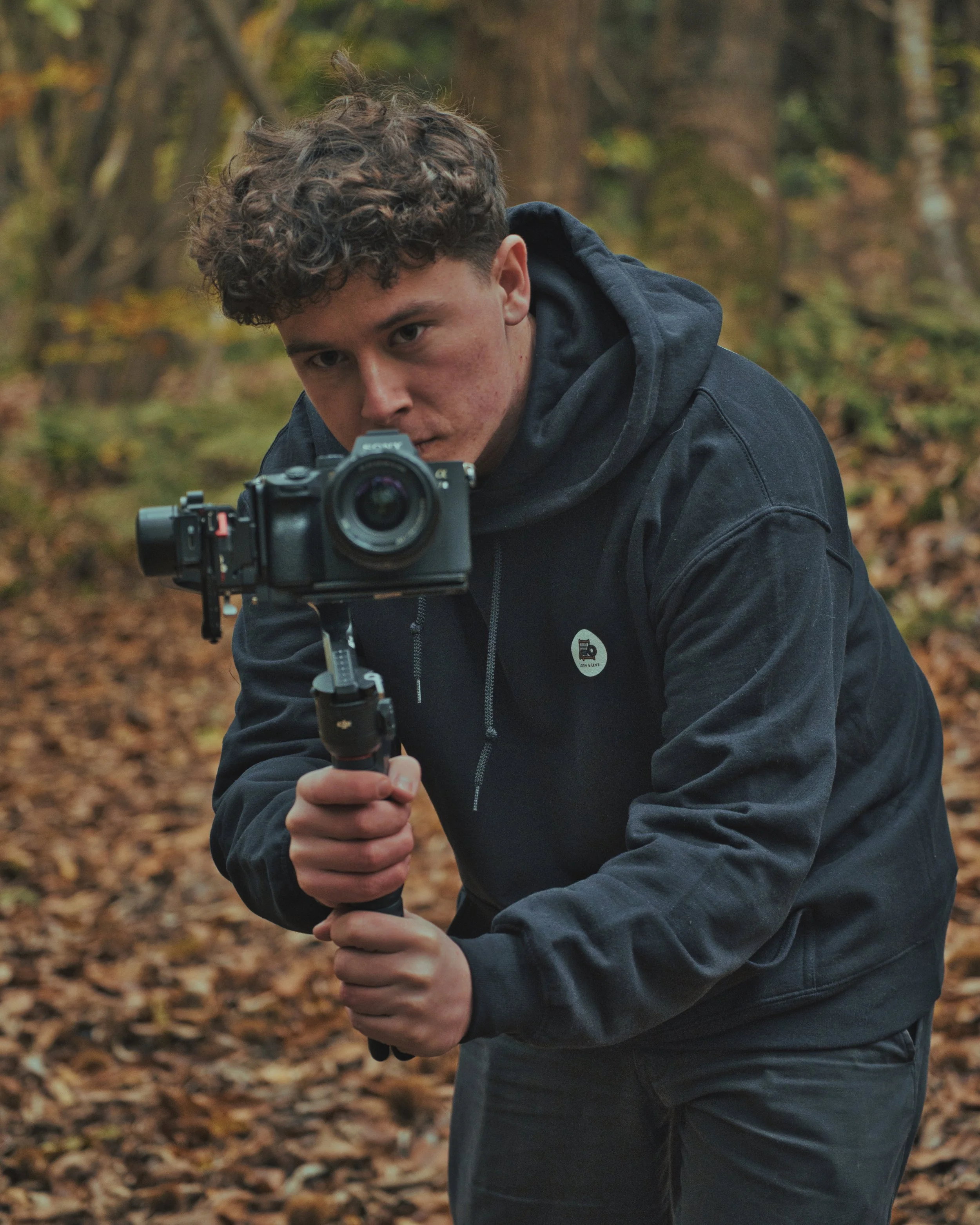 A young man with brown curly hair in a black hoodie holding a camera mounted on a stabilizer, filming in a forest with fallen leaves on the ground.