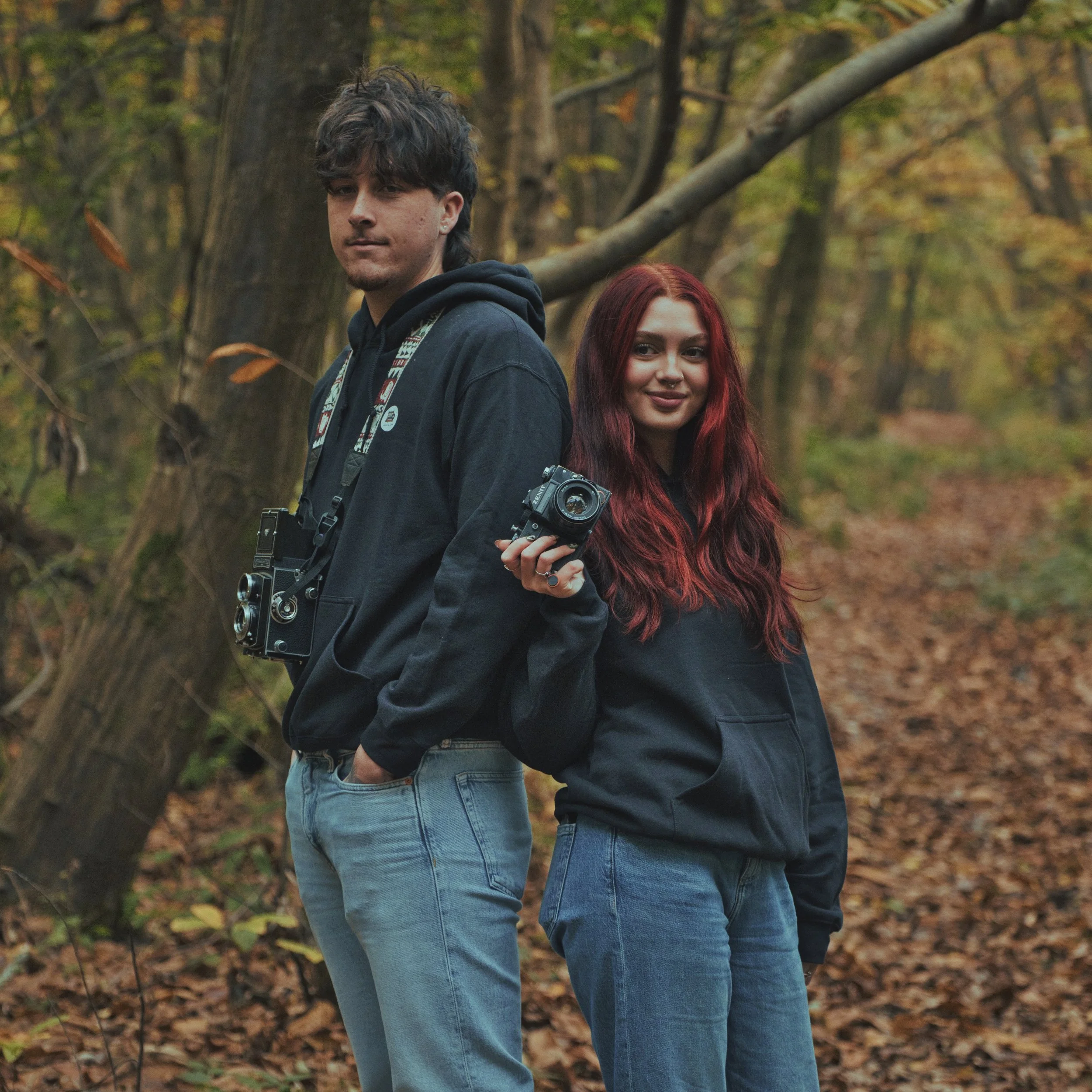 A young man and woman stand back-to-back in a wooded area during autumn, both holding cameras and smiling.
