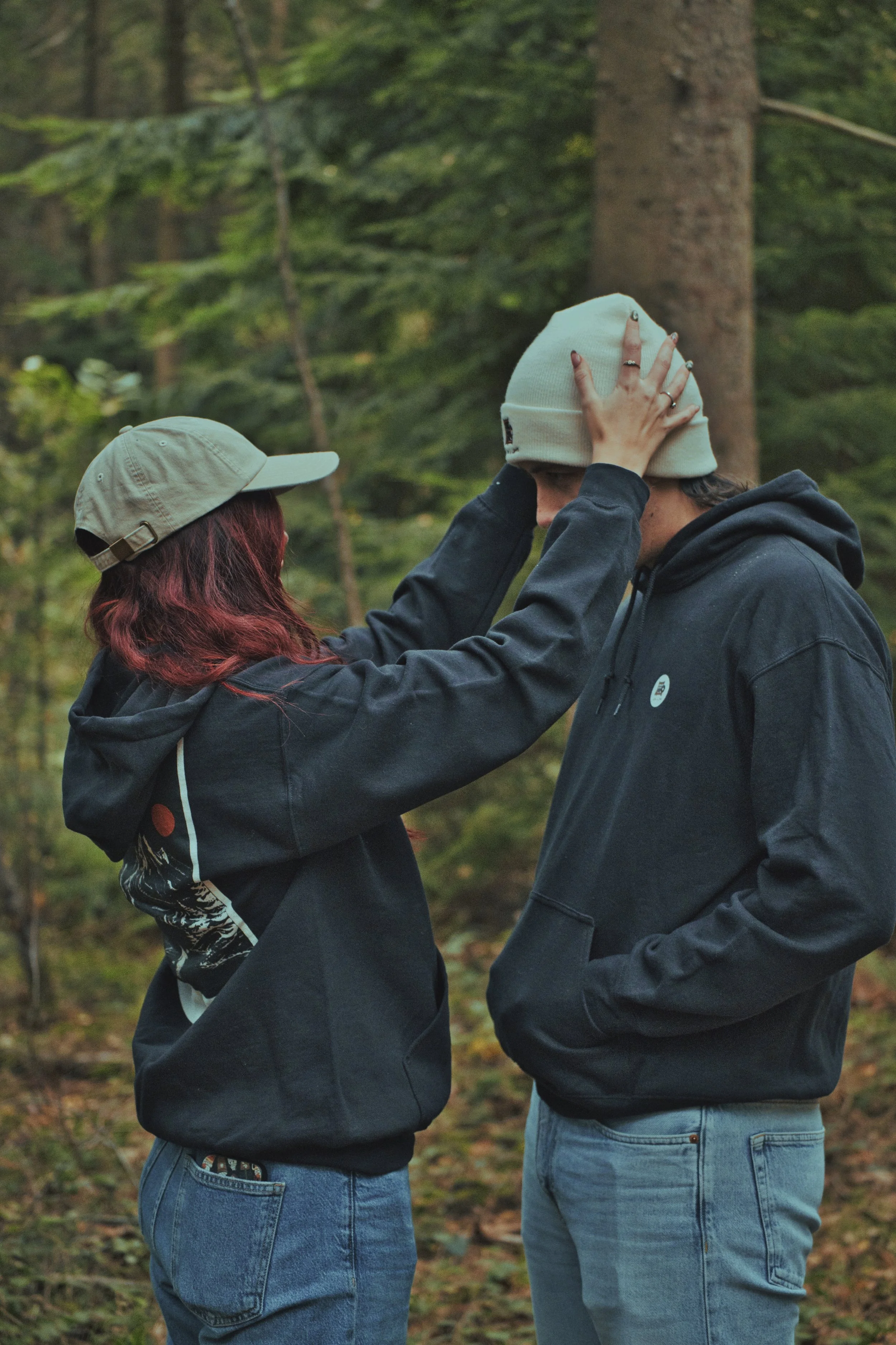 A woman with red hair and a beige baseball cap is touching the face of a man with a dark hoodie and a white beanie in a forest.