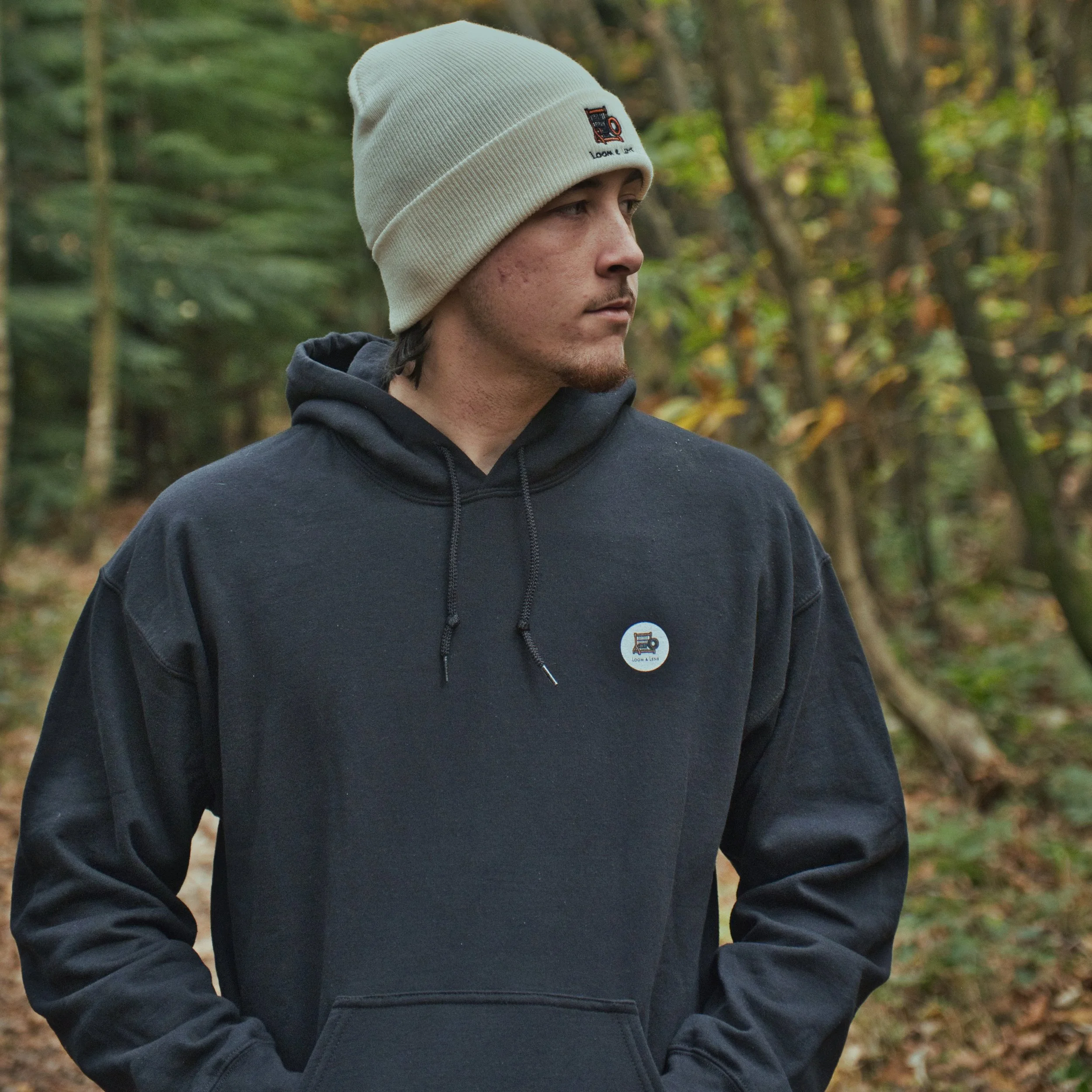 Young man wearing a beige knit beanie and black hoodie with a small logo pin, standing outdoors in a wooded area with trees and fallen leaves.