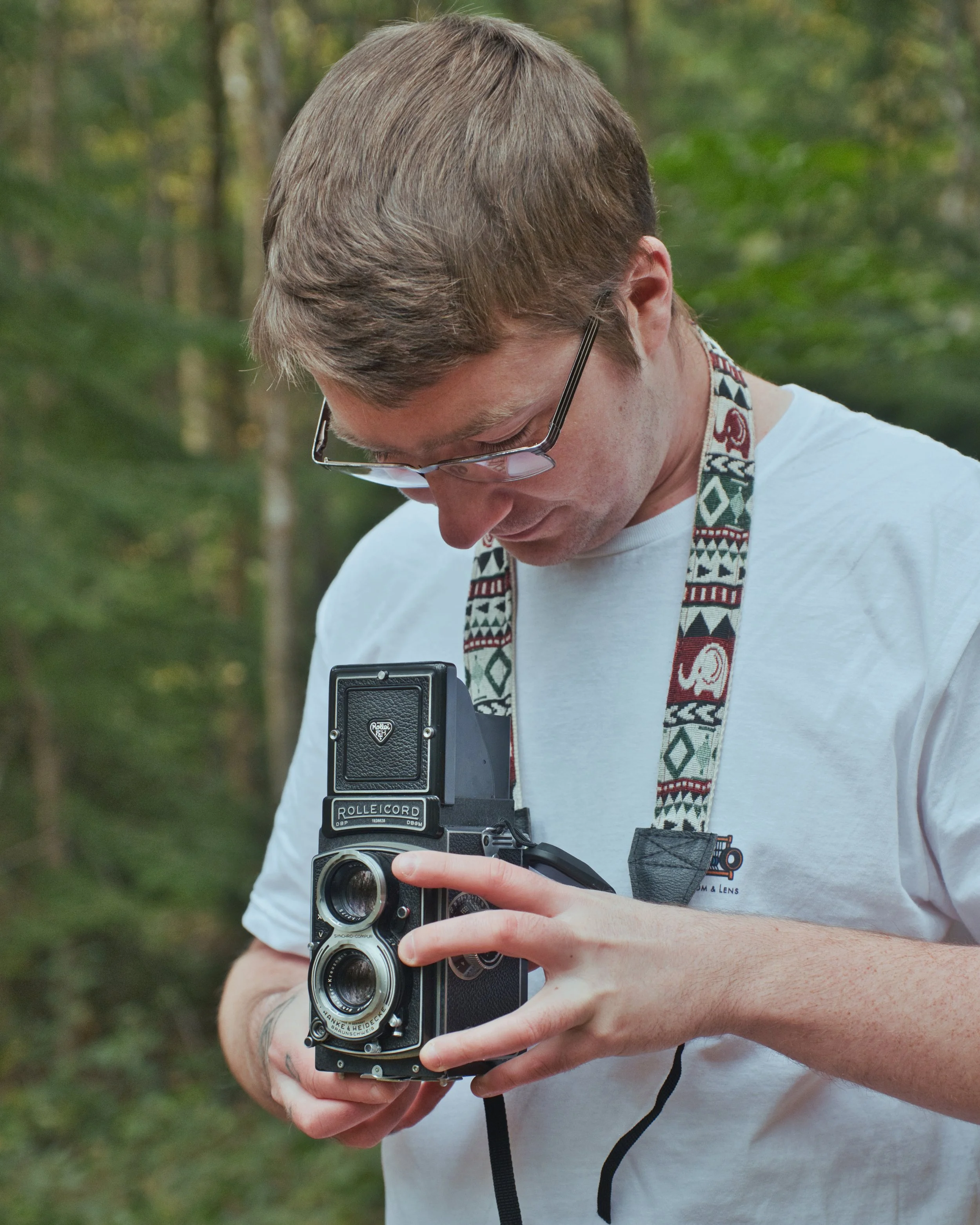 A man with glasses and a patterned camera strap looks down at a vintage twin-lens reflex camera while outdoors in a wooded area.