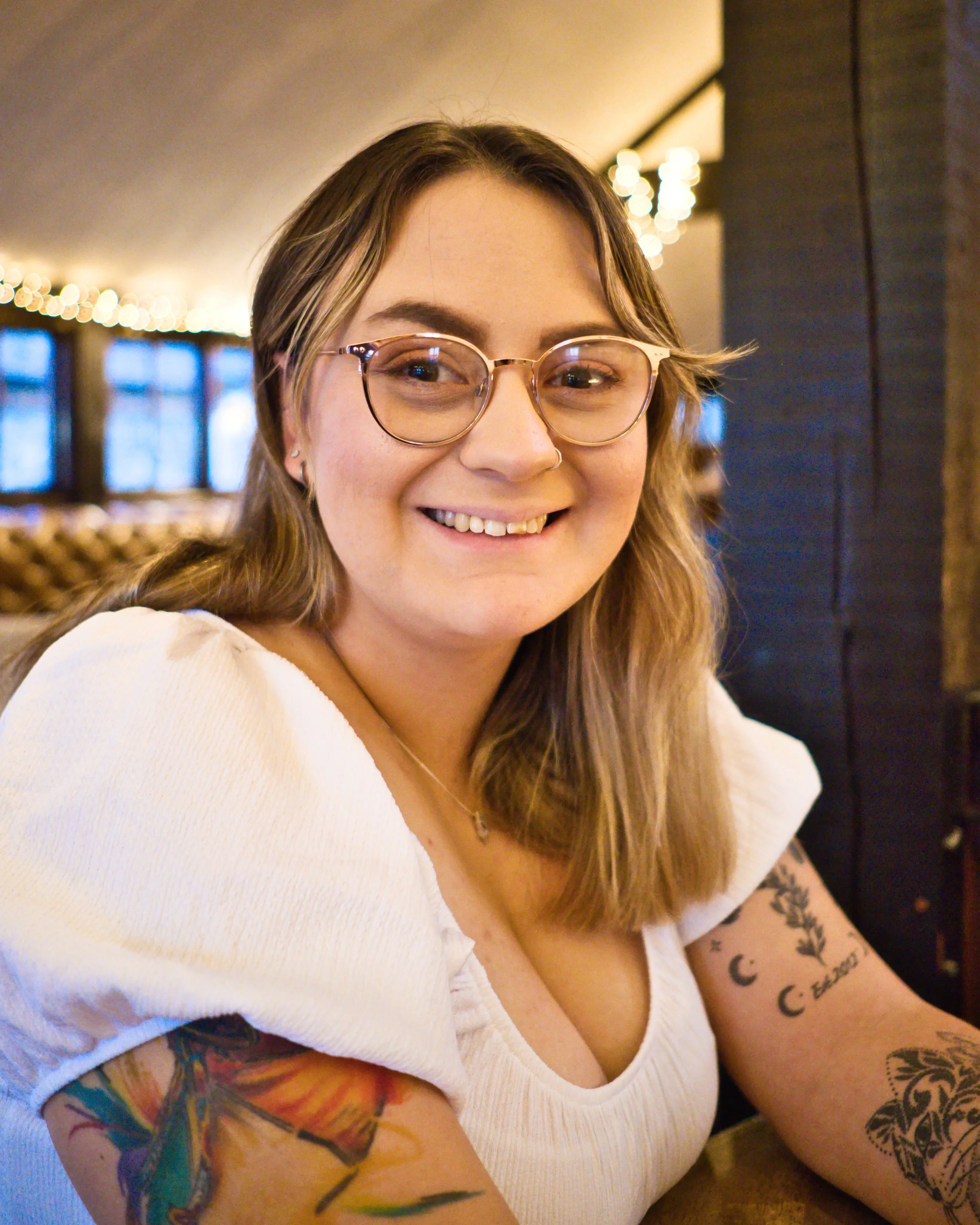 A young woman with glasses, tattoos, and a nose piercing sitting indoors, smiling at the camera.