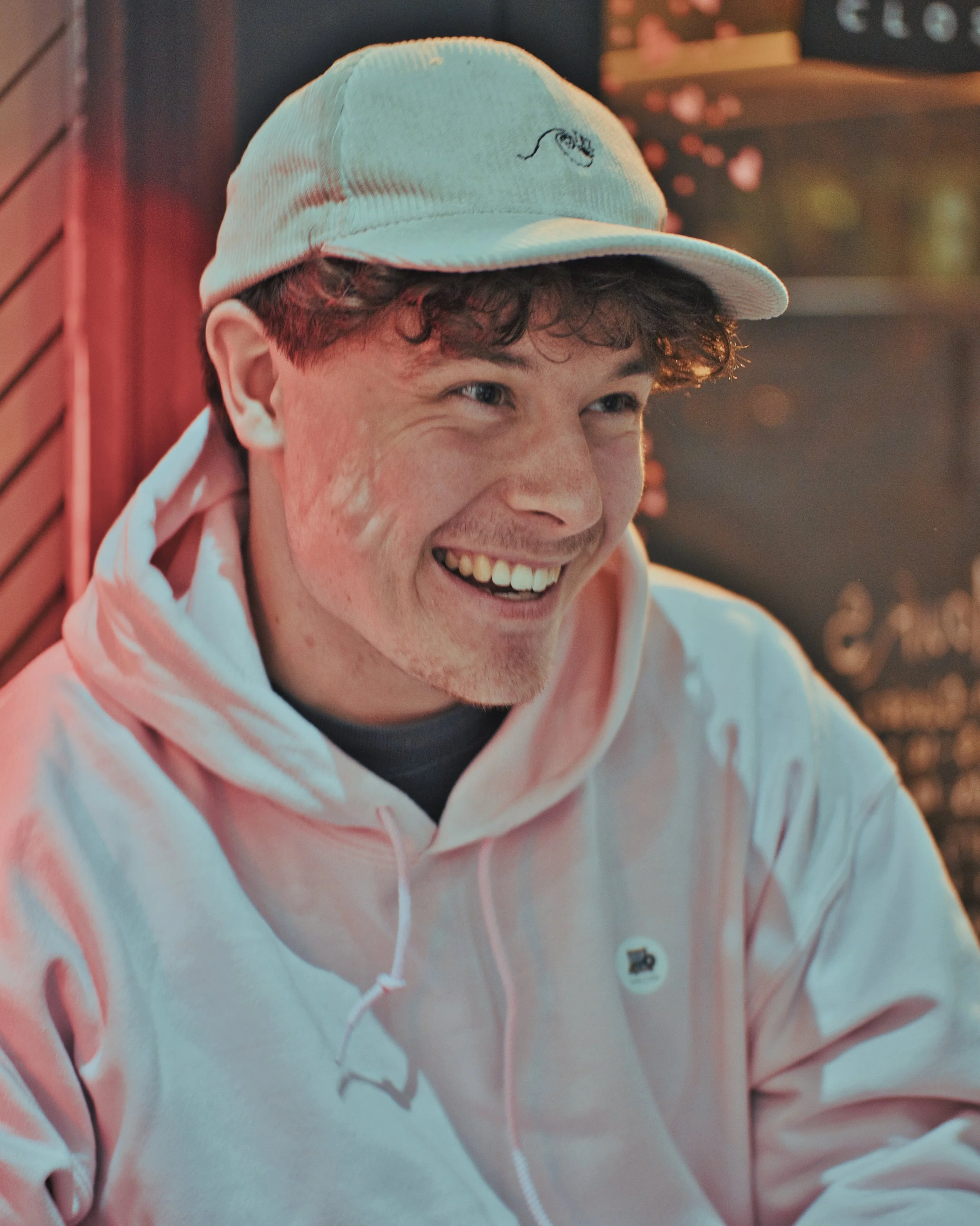 Young man smiling, wearing a light-colored hoodie and a beige cap with a loom and lens logo embroidered on it, sitting indoors with warm lighting.